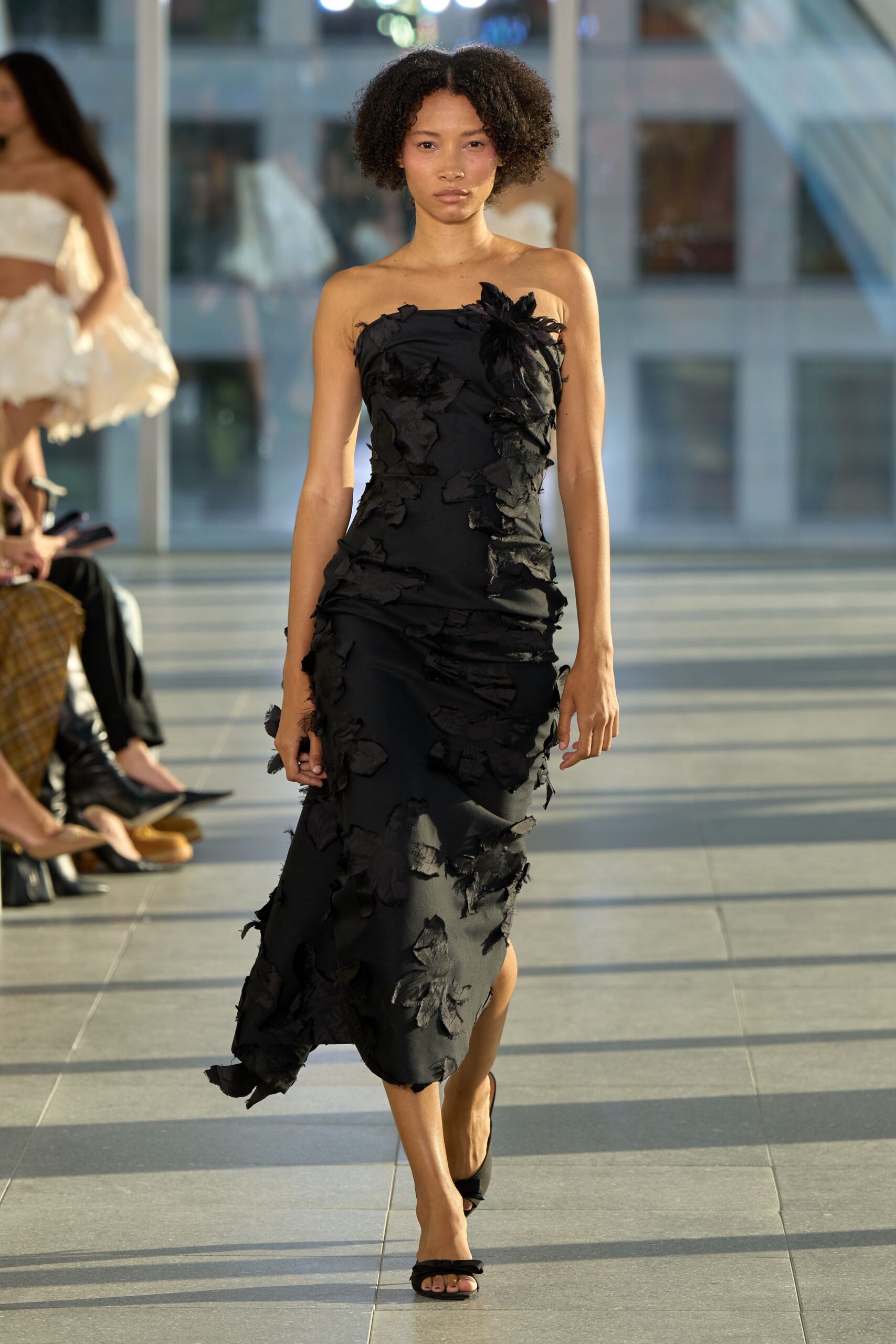 Black column dress appliqued with frayed petal shapes, one clustering at the bust. Black satin slide sandals with a narrow twisted vamp. Crossing a glass atrium when the sun drops low enough to flatten everything.
