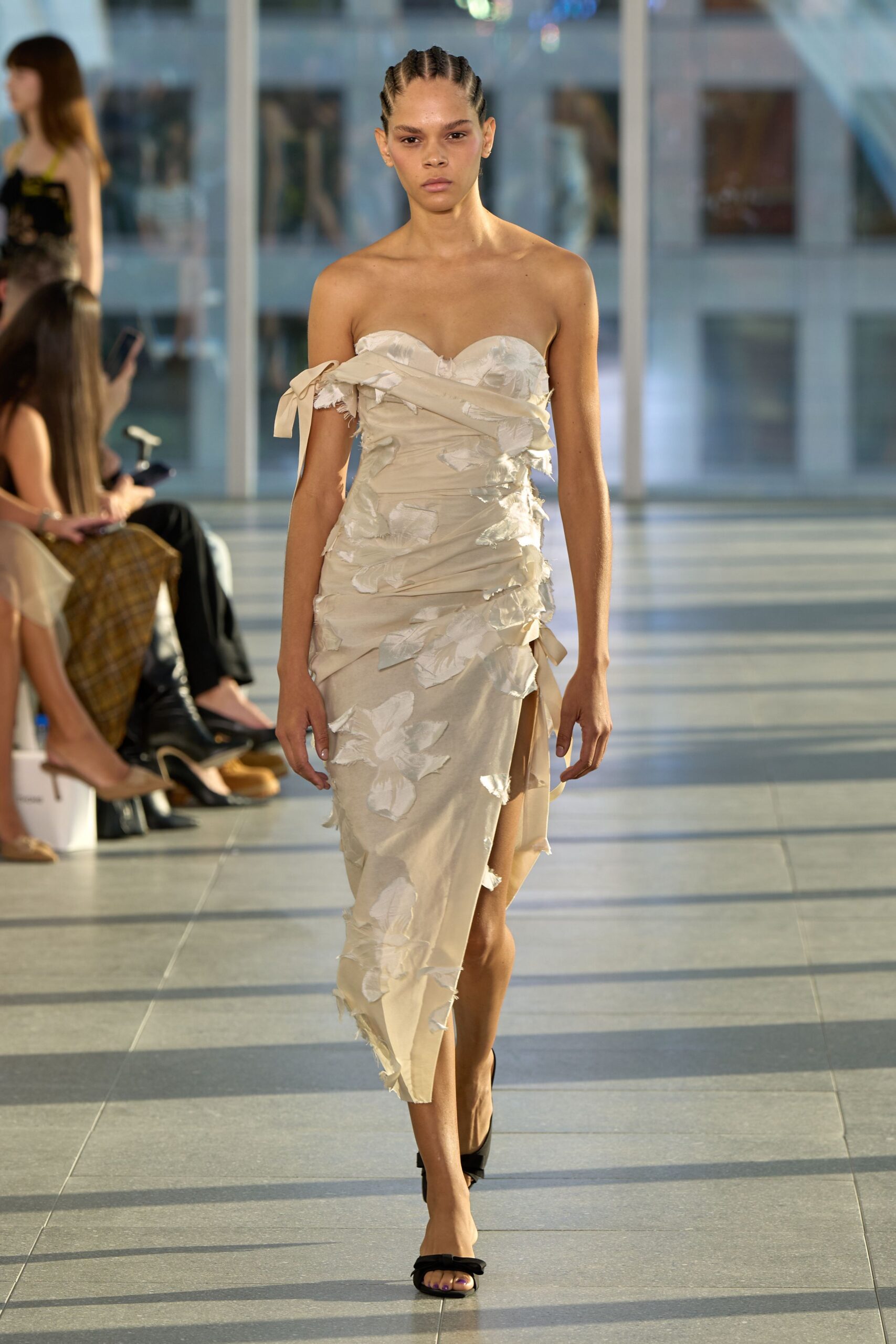 Strapless sand satin dress ruched tight, with frayed petal appliqués and a tied off-shoulder band. Black bow-front sandals with a narrow bare thong. Walking through a glass atrium after someone else's wedding has ended.
