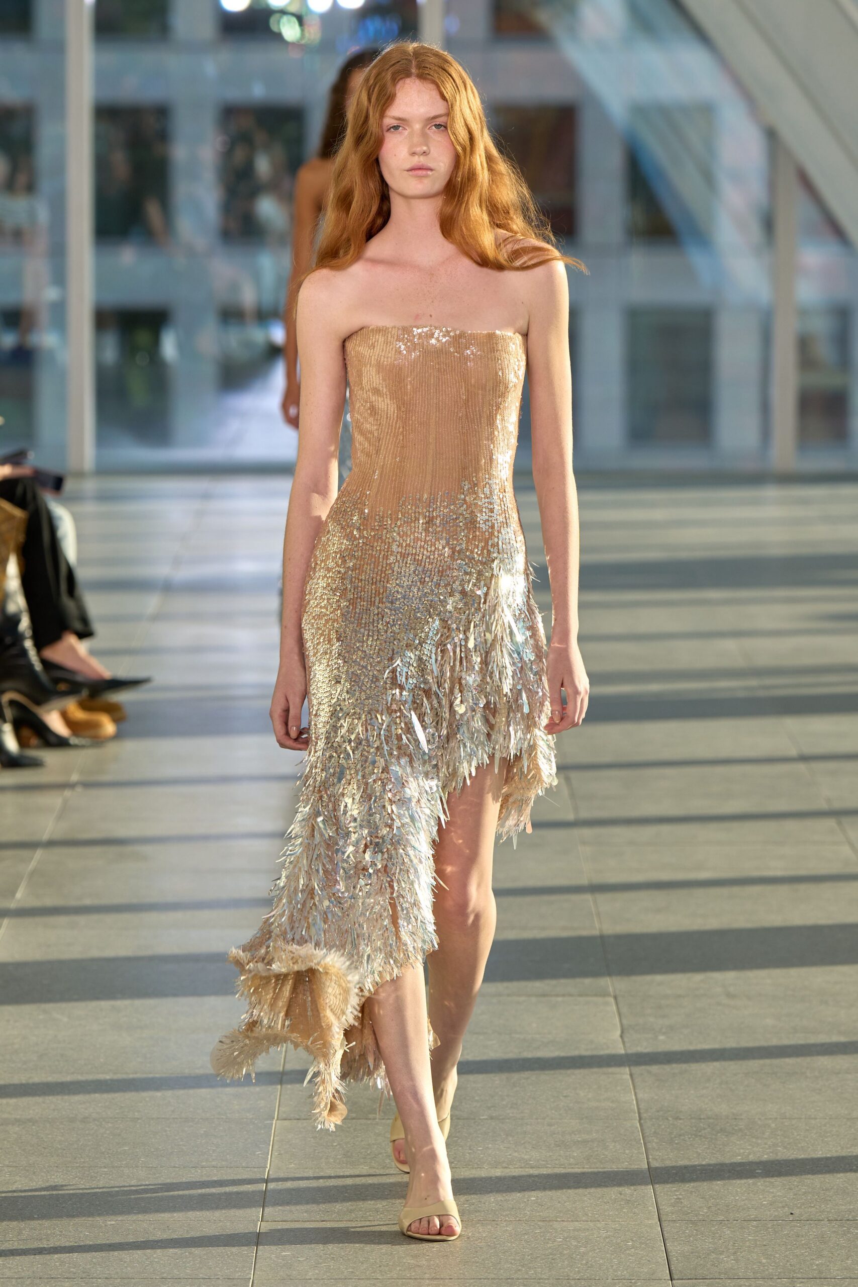 Strapless champagne column densely beaded, dissolving into an iridescent feathered train. Nude slide sandals with a squared toe. Crossing a glass atrium just as the low sun turns everything to metal.