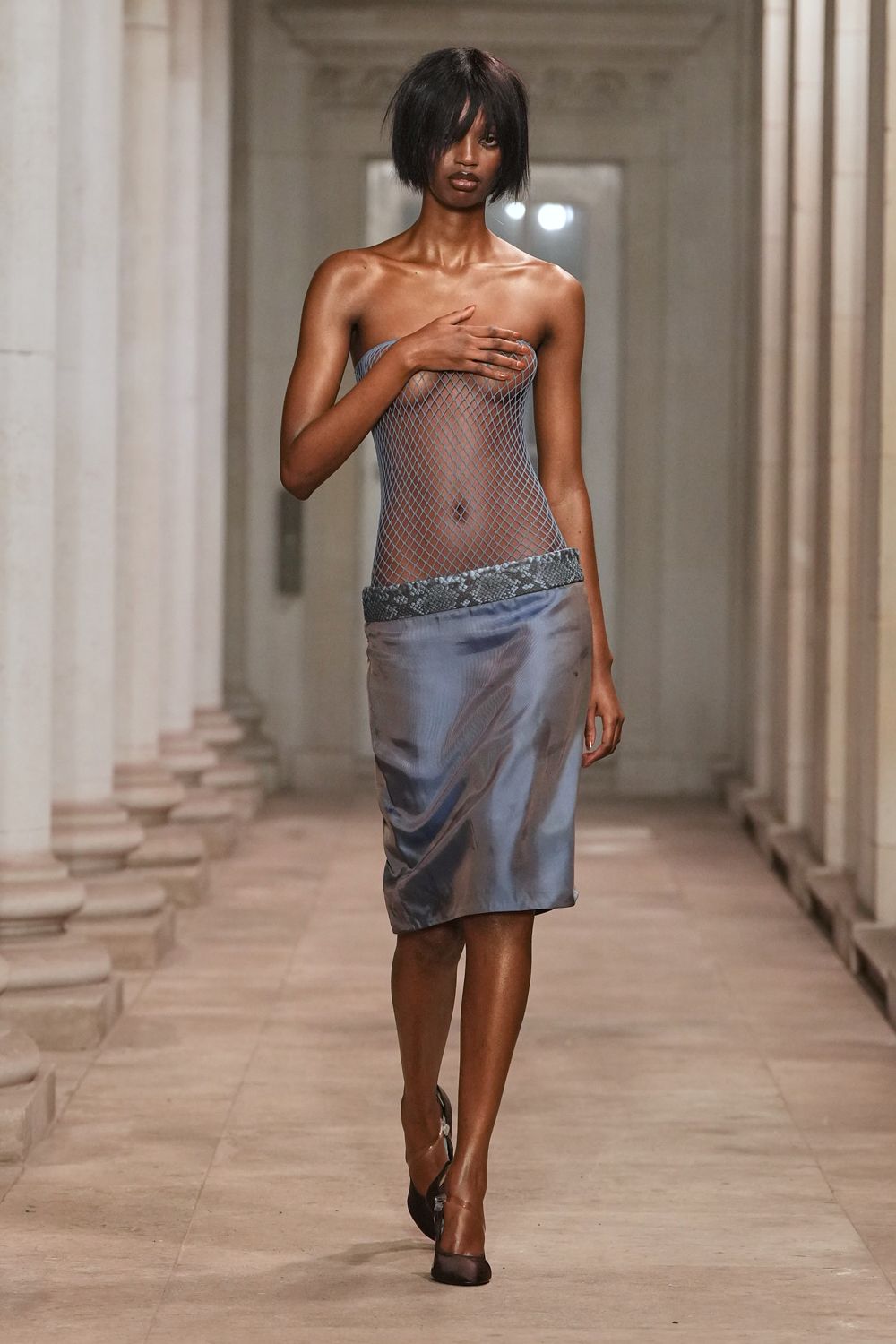 Sheer steel-blue fishnet tube, stretched wide across the bust. Blue-grey satin pencil skirt, low slung under a python band. Black mesh pumps with a thin ankle strap. Whatever you wear home after the hotel pool emptied at dusk.