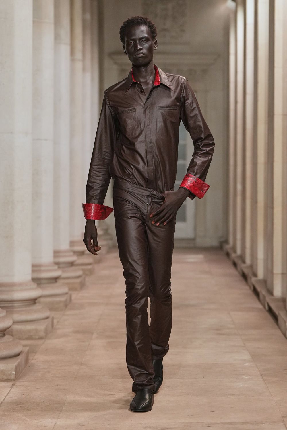 Chocolate leather shirt with a concealed placket and scarlet python cuffs. Matching leather trousers, low at the waist with a twisted overlap closure. Black square-toe ankle boots. Crossing a museum cloister after rain, still carrying the argument.