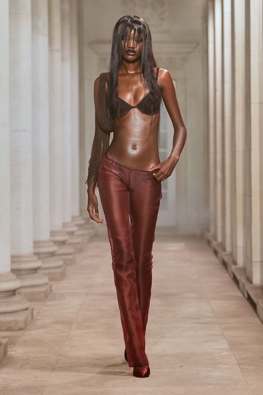 Dark brown bra with sharply arched cups and thin straps. Sheer brown shoulder bag finished with long fringe. Burgundy sheer trousers, cut low and skimming over the shoes. Pointed satin pumps in the same wine shade. Crossing a cloister courtyard after midnight rain, stone still holding the heat.
