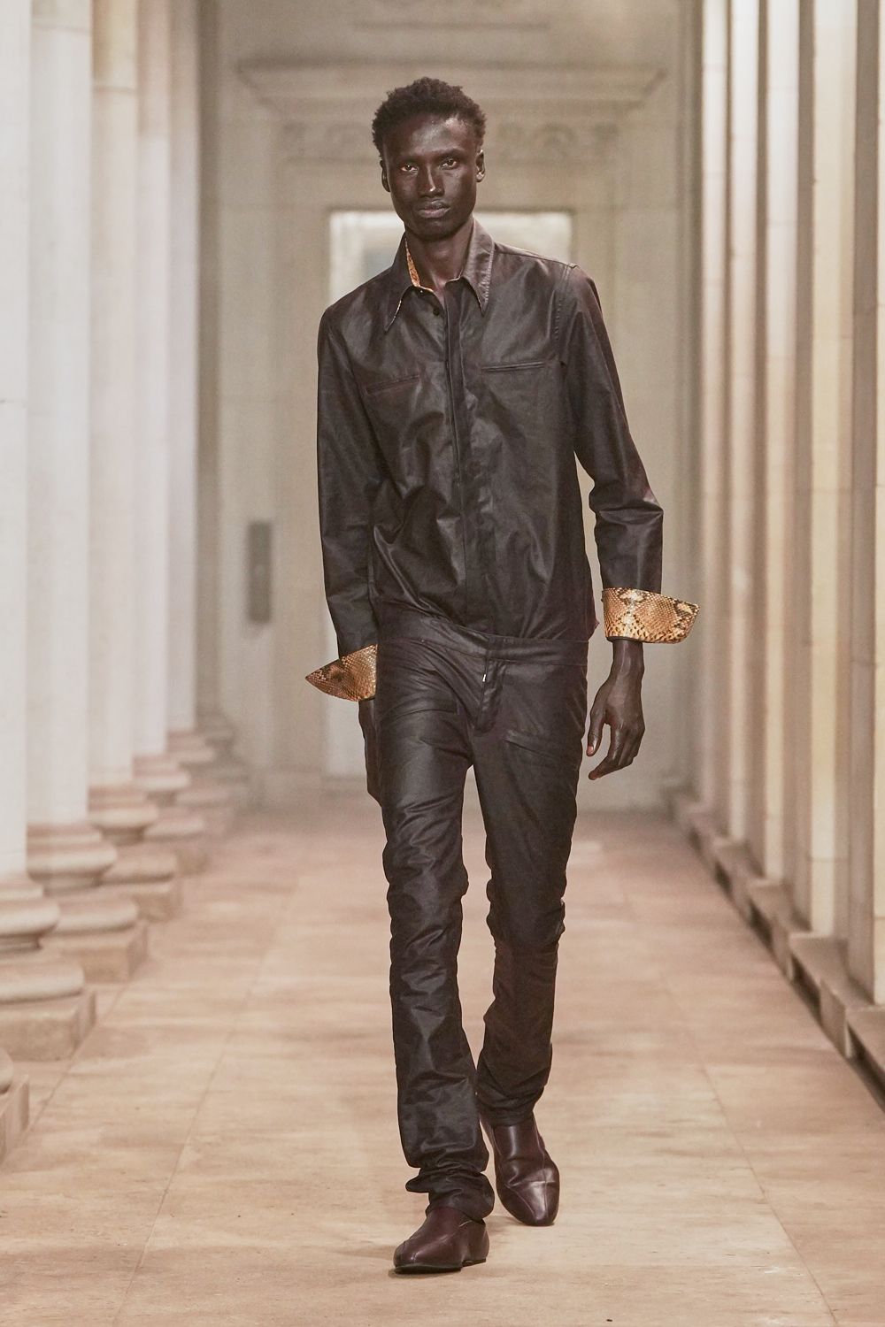 Black coated shirt with flap pockets and turned-back python cuffs. Low-slung black coated trousers, zip fly sitting slightly open. Dark square-toe leather boots. Crossing a museum corridor before the public doors unlock.