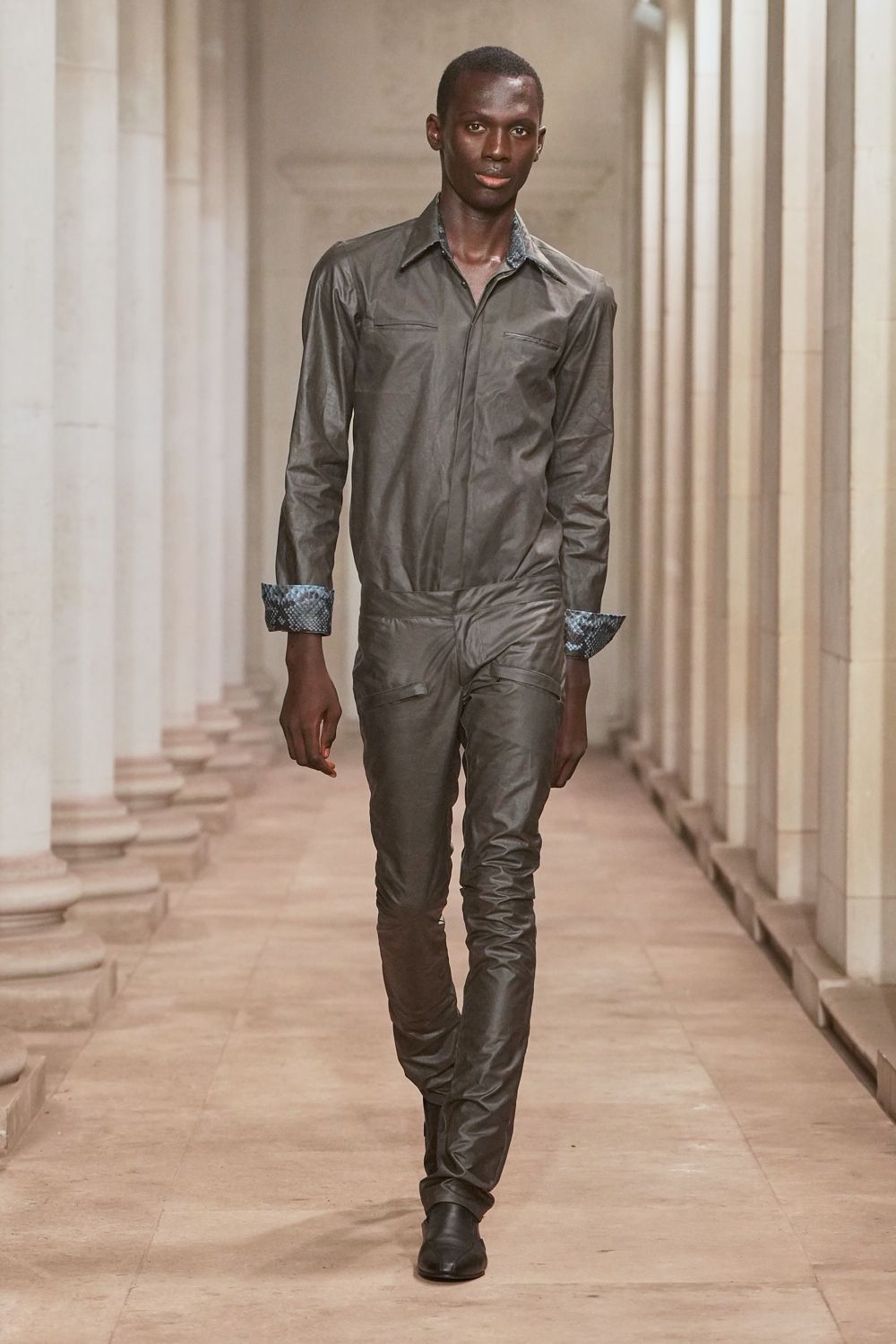 Slate leather jumpsuit with zipped chest welt pockets and a dropped waist. Turned-back cuffs lined in blue-grey python print. Black square-toe ankle boots, nearly swallowed by the narrow leg. Crossing a museum cloister after rain, keys still warm in your hand.