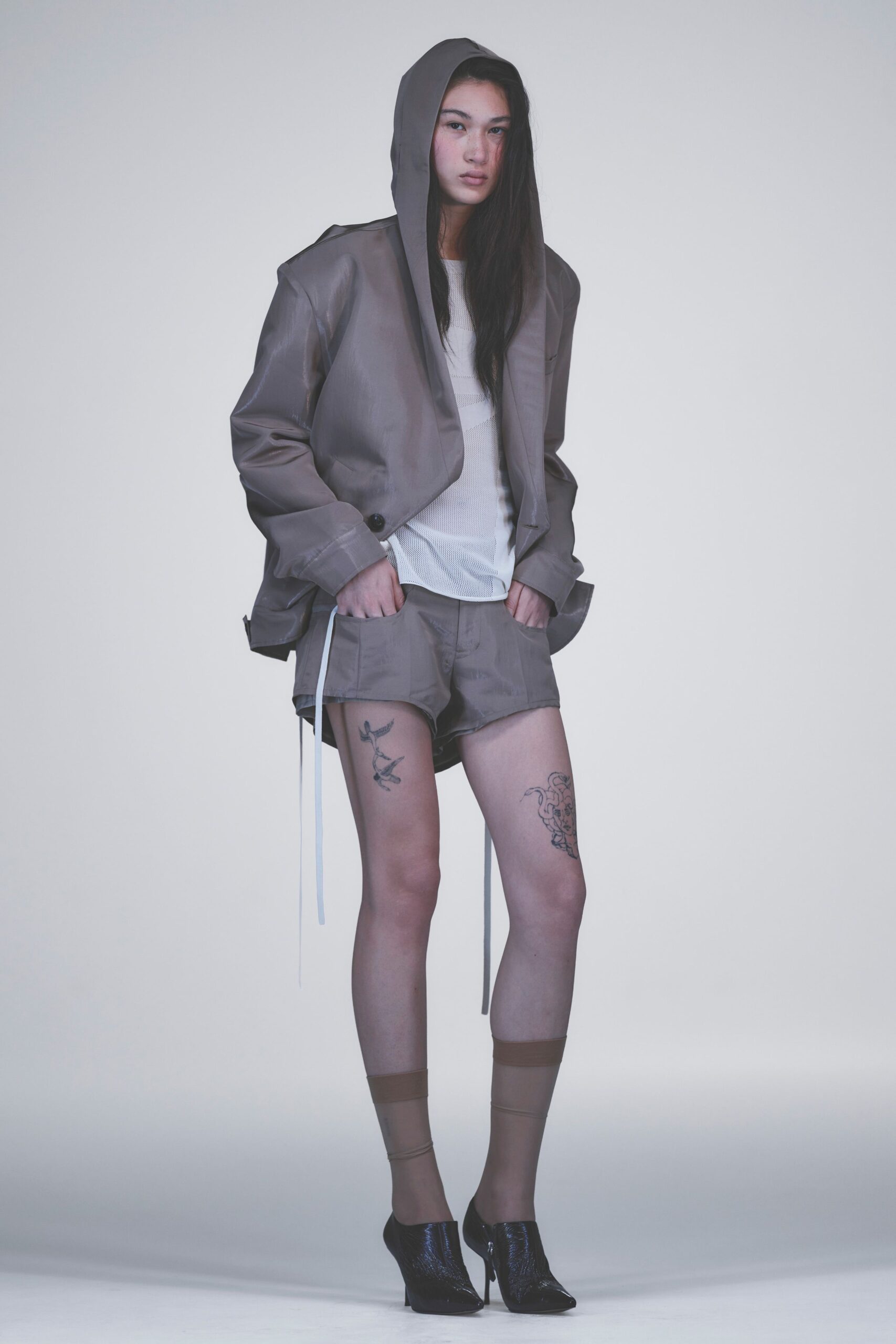 Taupe hooded jacket in crisp coated cloth, cut with a single dark button. White open-knit top. Matching taupe shorts with long white drawcords hanging past the hem, sheer socks, black crinkled patent mules. Waiting in a rehearsal studio before dawn.