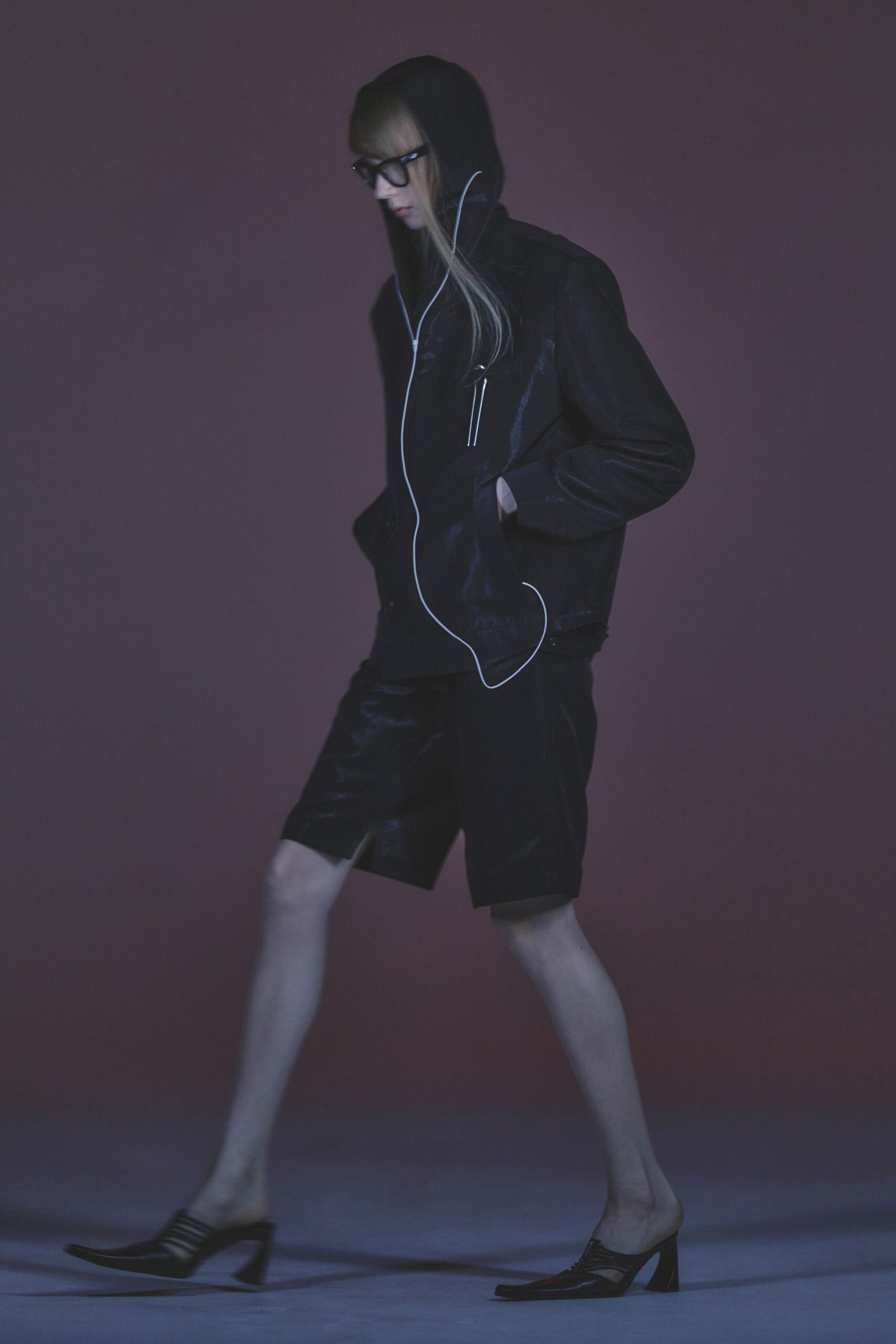 Thick black rectangular frames. Black satin-finish hooded jacket with white zipper piping snaking off-center. Knee-length black shorts, one leg catching a vinyl gloss. Pointed black mules with cutout sides and stacked heels. Walking to the corner shop after midnight with earbuds still in.