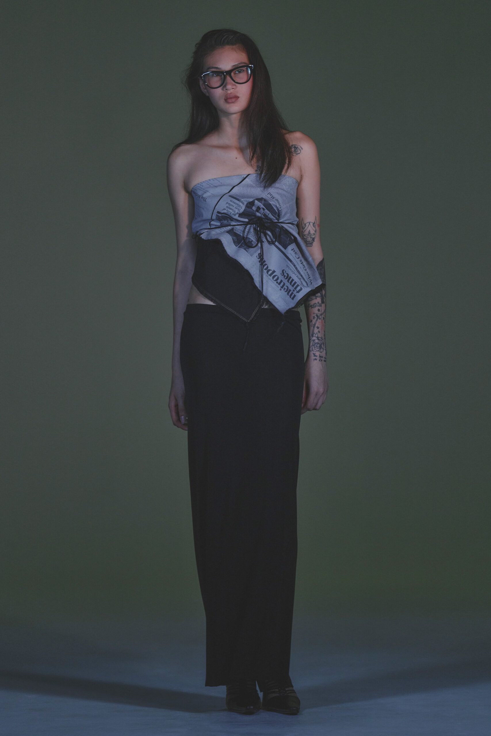 Thick black acetate frames catching a hard white edge. Newspaper-print scarf top, black cord laced tight across the bust. Long black skirt hanging straight from a dropped, off-center waist. Square-toe black boots. Walking home from the corner store with the paper tucked under one arm.