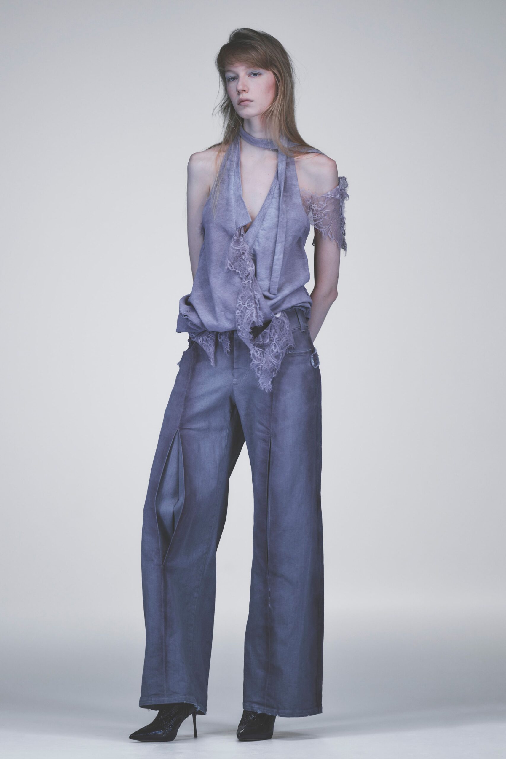 Lavender draped halter top with eyelash-lace panels trailing from the plunge and one shoulder, the neck strap looped close like a scarf. Washed slate wide-leg trousers with pressed creases, black patent ankle boots. Dawn in an empty rehearsal studio.