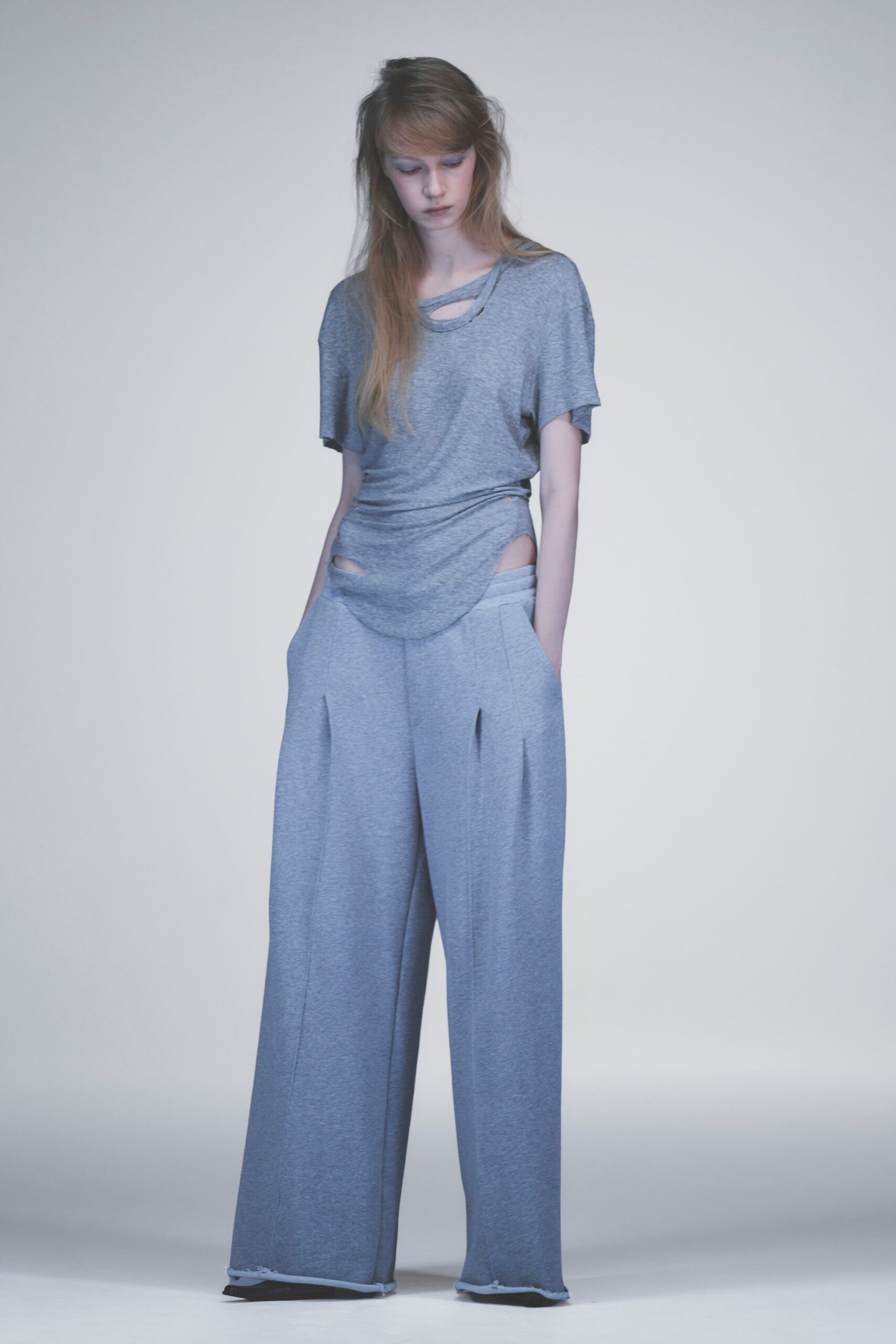 Heather-grey jersey tee with a slashed crewneck and skewed side drape. Matching marl knit wide-legs, cutout hips and raw hems curling above the floor. Whatever you wear home when daylight hits the after-hours grocery.