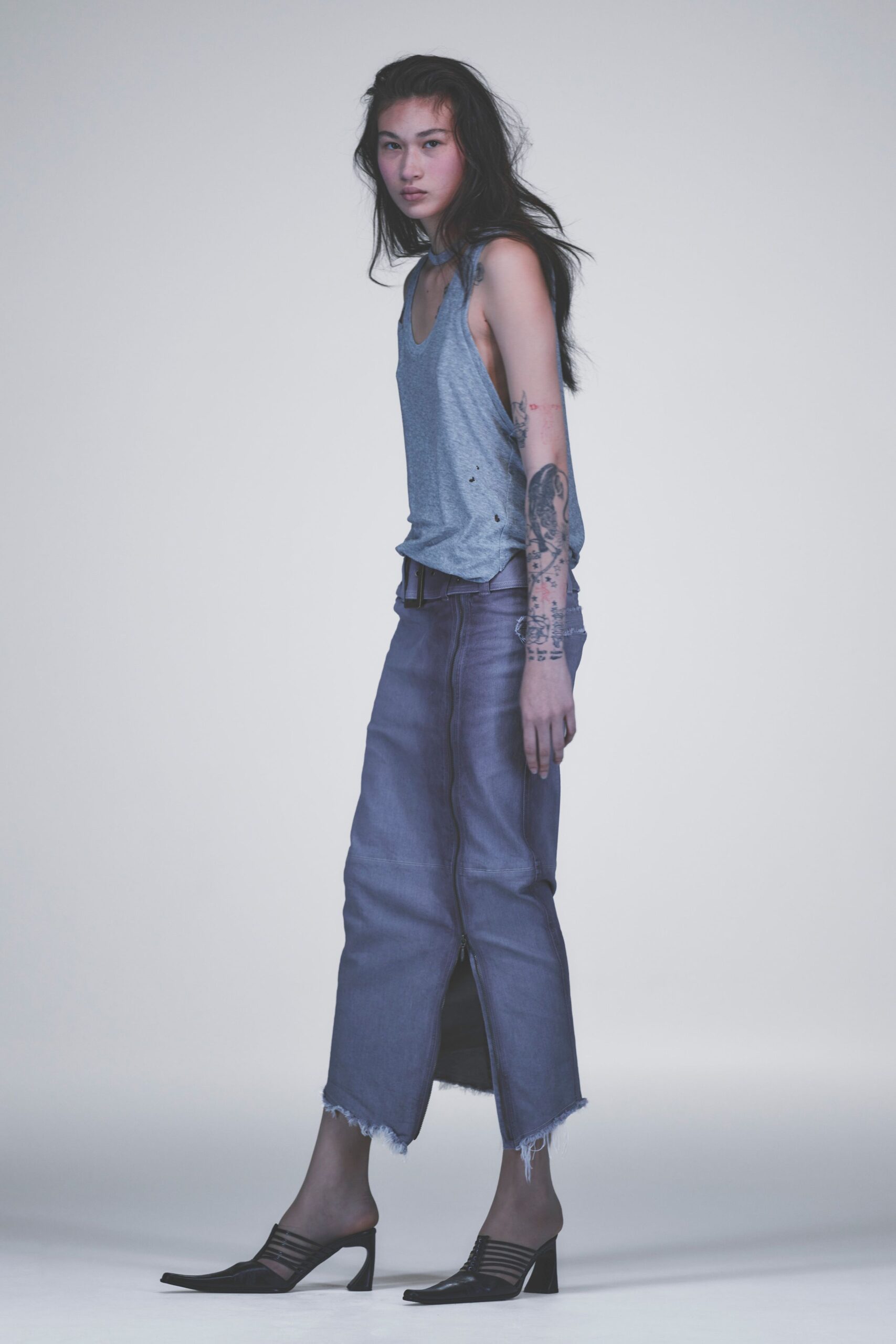 Heather-grey jersey tank with dropped armholes and a narrow neck band. Black leather belt with a plain rectangular buckle. Washed slate denim tube skirt, front zip running nearly to the hem. Black leather mules with caged straps and a squared pointed toe. For buying cigarettes at dawn with last night's eyeliner still on.