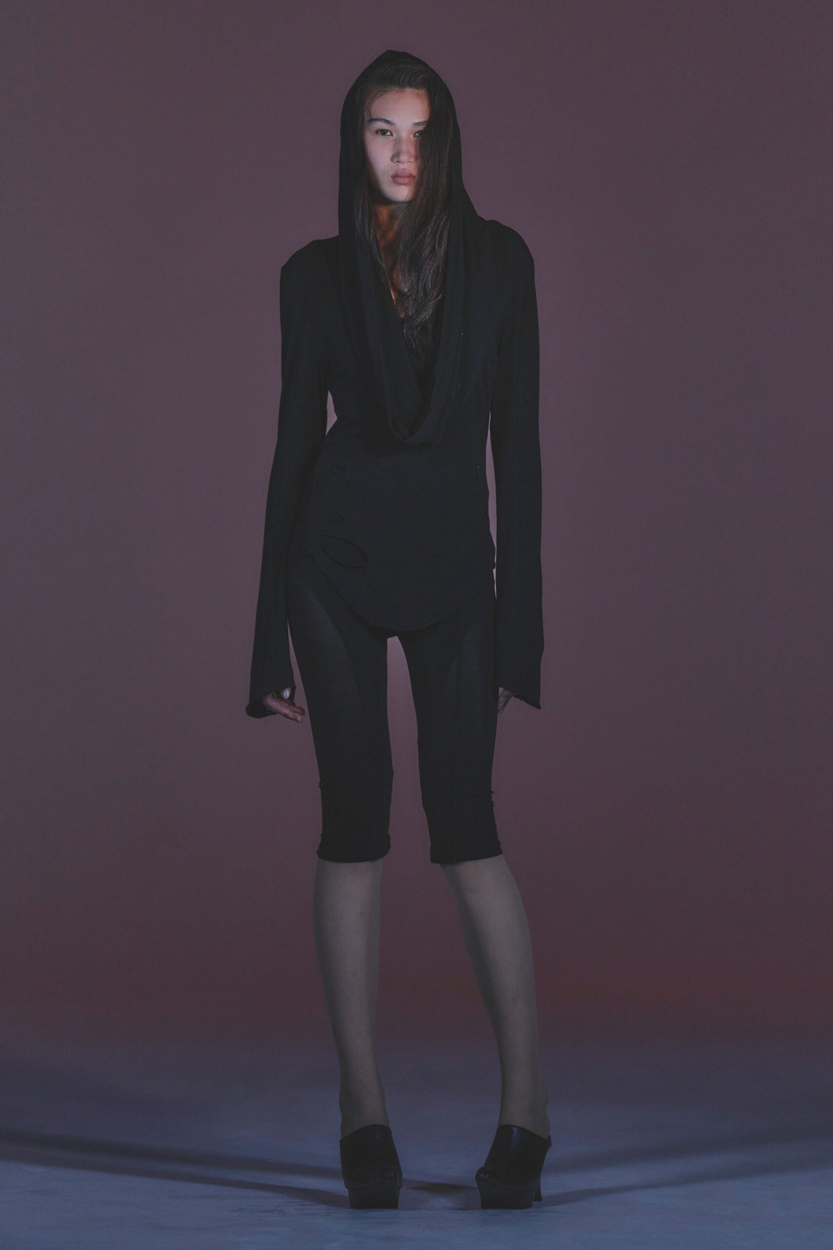 Black jersey hooded top with a deep cowl plunging past the sternum. Matte black cropped leggings, ending tight just below the knee. Black leather platform mules with a blunt square toe. Walking home after the club, dawn turning the side streets violet.