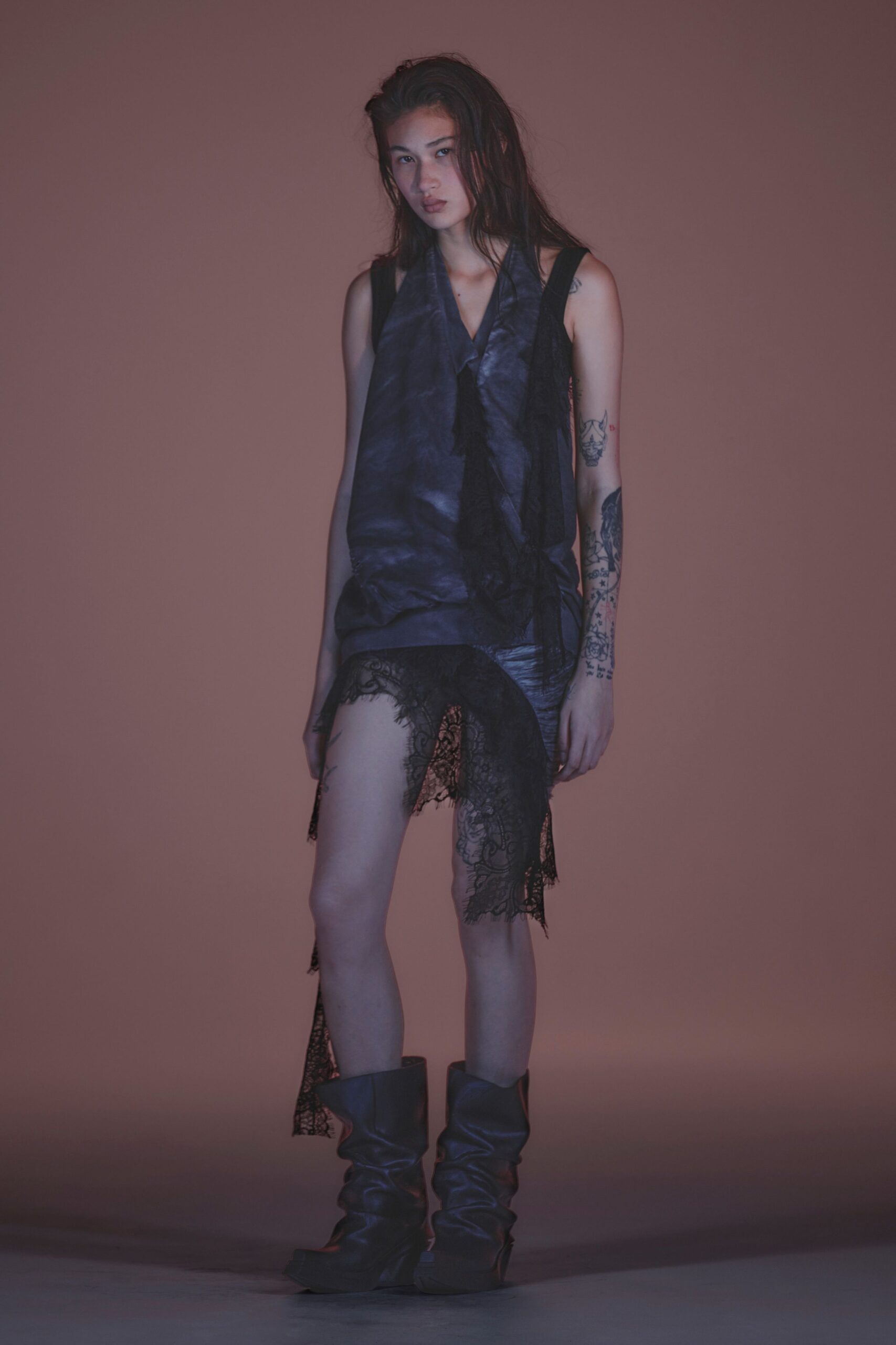 Ink-dark satin tunic with a draped scarf front and hanging ties. Black eyelash lace skirt, one panel dropping nearly to the ankle. Slate leather platform boots slouched into heavy folds at the shin. After midnight in a karaoke booth, one heel stuck to the vinyl floor.