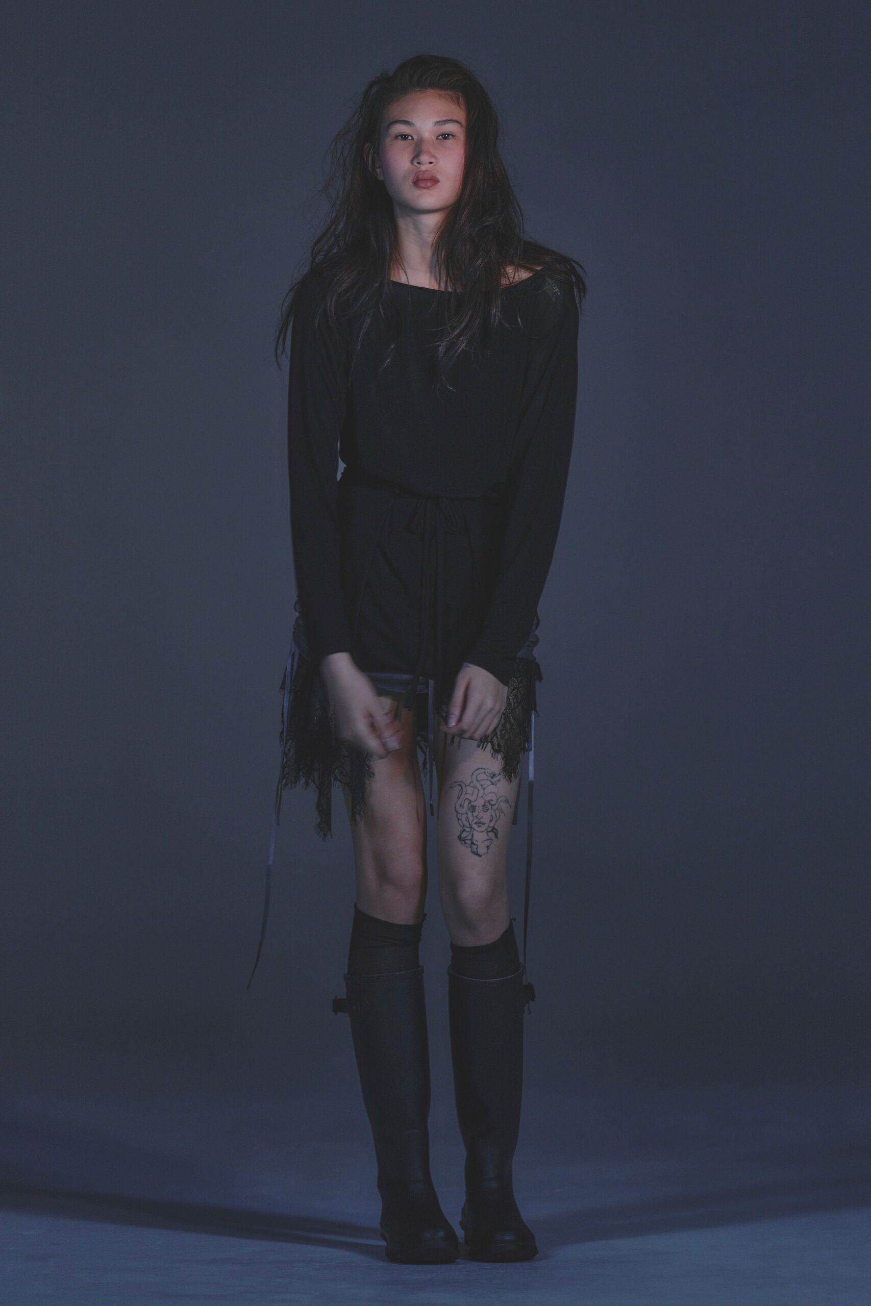 Black jersey tunic with a boat neck slipping off one shoulder. Black mini skirt with lace panels and long dangling drawcords. Black slouch socks. Matte black knee boots with a side buckle near the calf. After midnight in a practice studio, tape marks still on the floor.