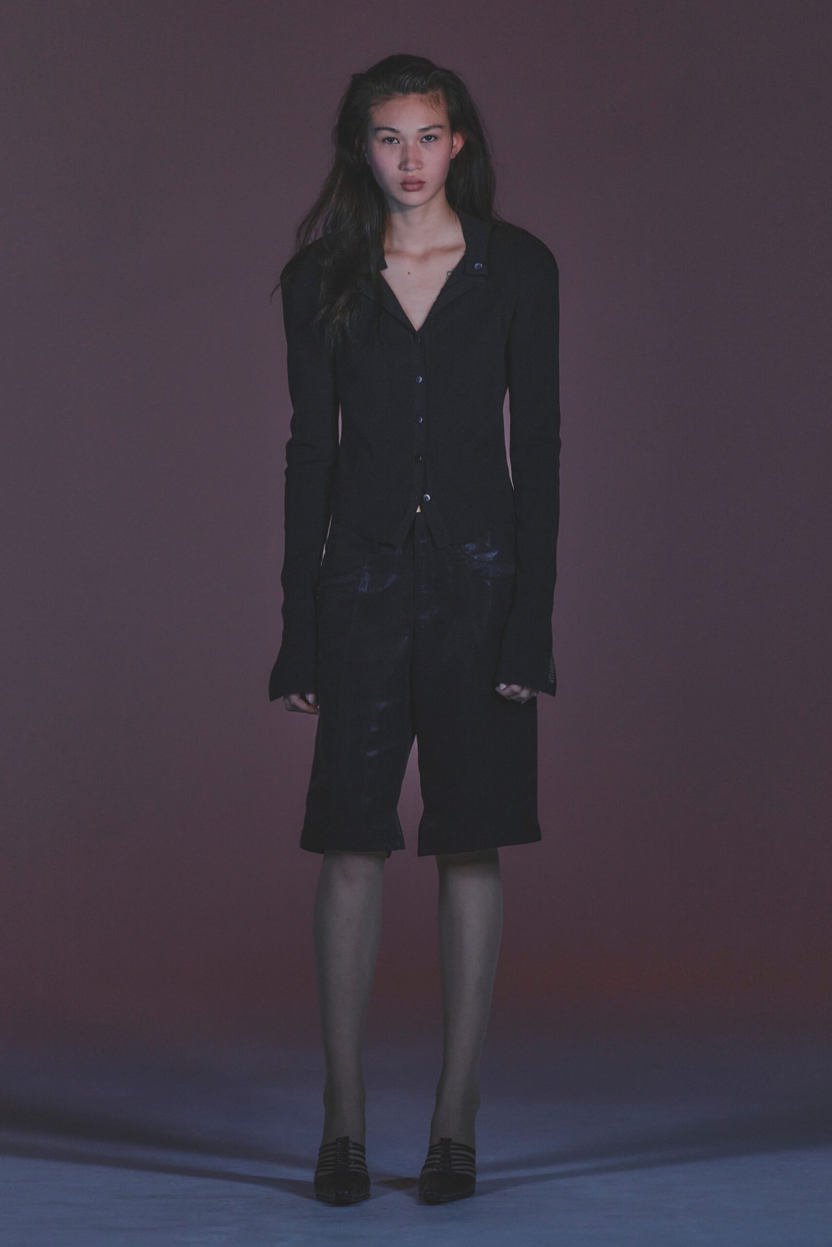 Black fine-rib cardigan with a spread collar and tiny blue-black buttons. Inky knee-length shorts with a waxy sheen catching across the front. Black cage-toe heels. After midnight at the hotel vending machine, ice bucket still in hand.