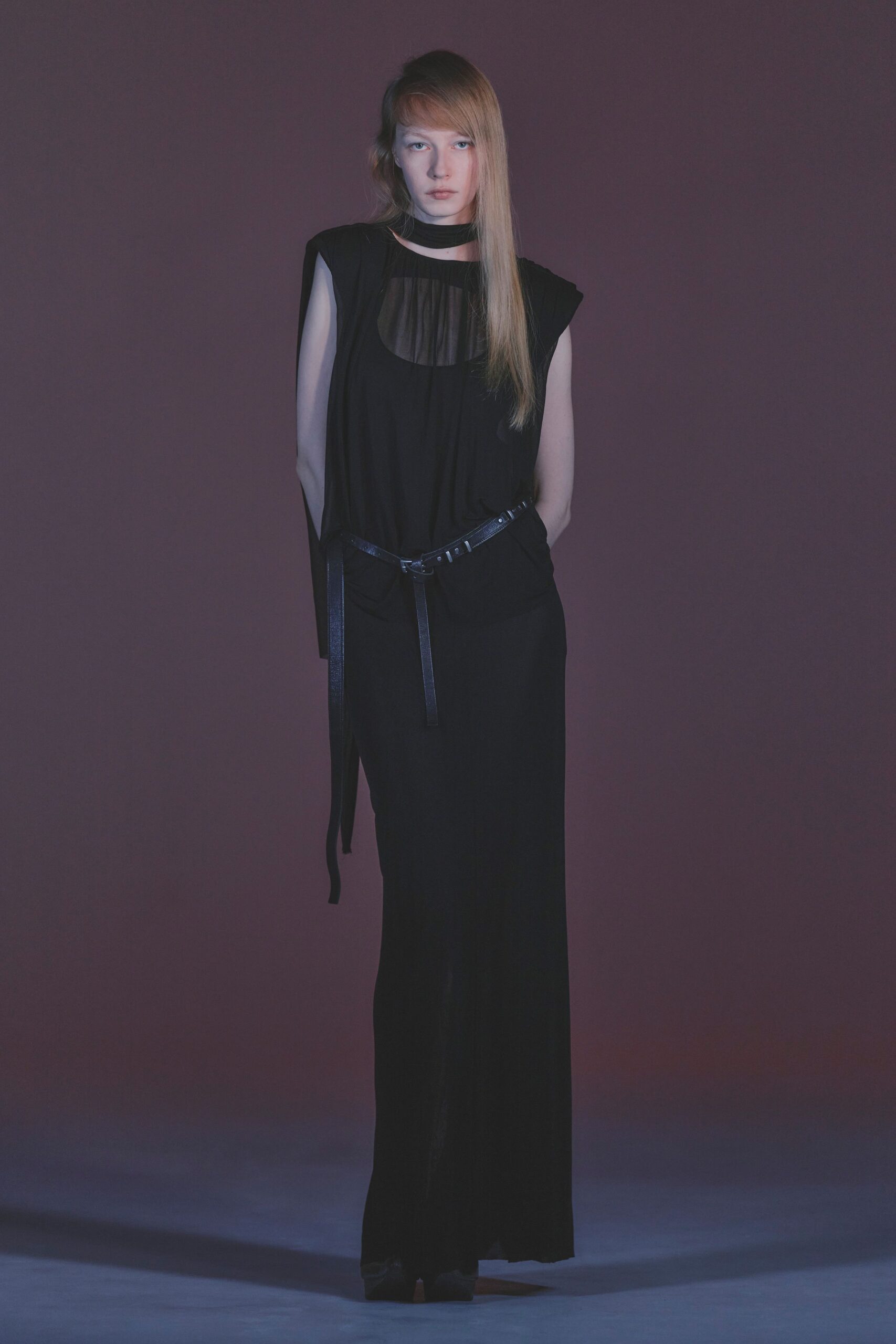Black draped jersey dress with a sheer oval chest inset. Narrow self-fabric band circling the throat, cut open at front. Thin black leather belt slung low, one strap hanging to the knee. Black square-toe shoes peeking from the pooling hem. After midnight in a hotel corridor, keycard demagnetized again.