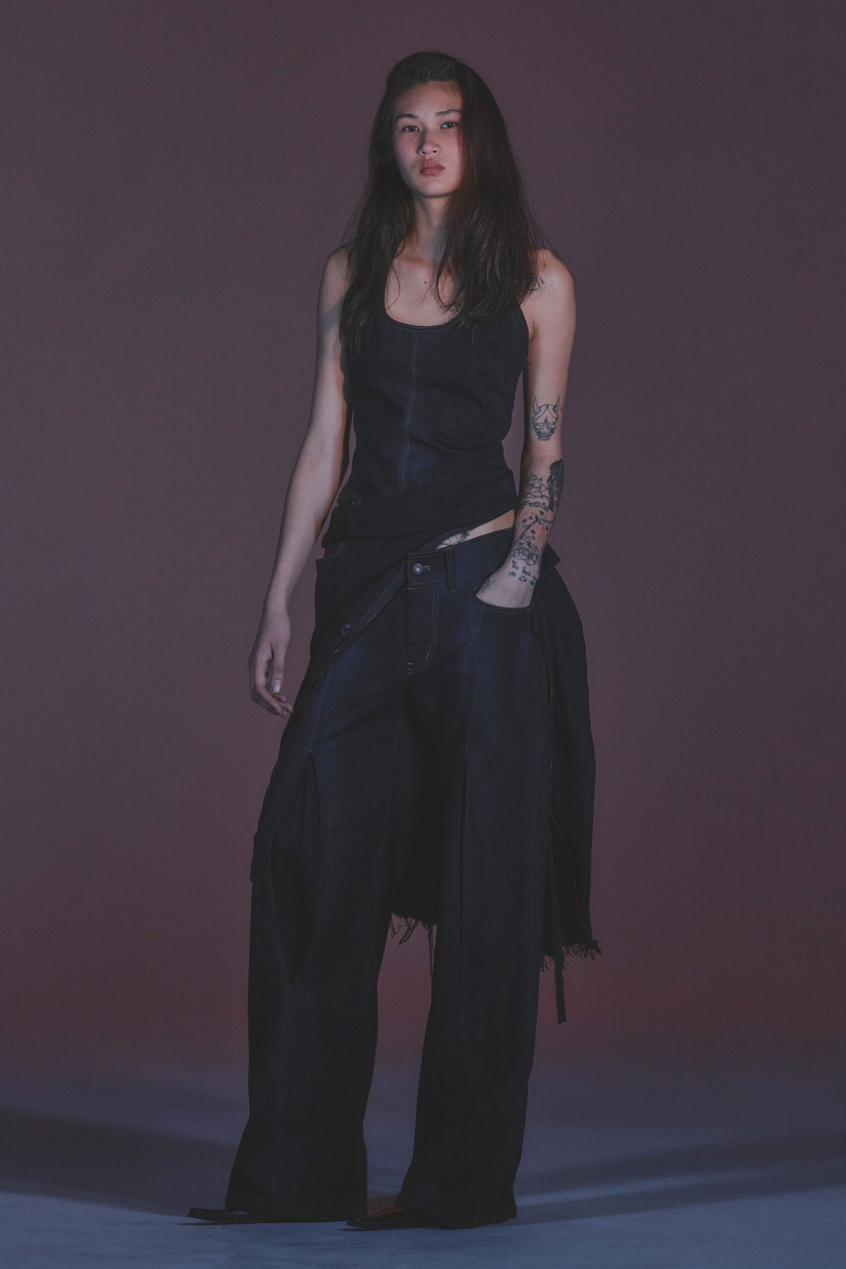 Inky fitted tank with a curved scoop neck and center seam. Black denim overskirt hanging from the hips, one frayed panel trailing. Dark indigo wide-leg jeans with contrast topstitching and a low slung rise. Square-toe shoes barely visible under the hem. For crossing the river at dusk after missing the last gallery opening.