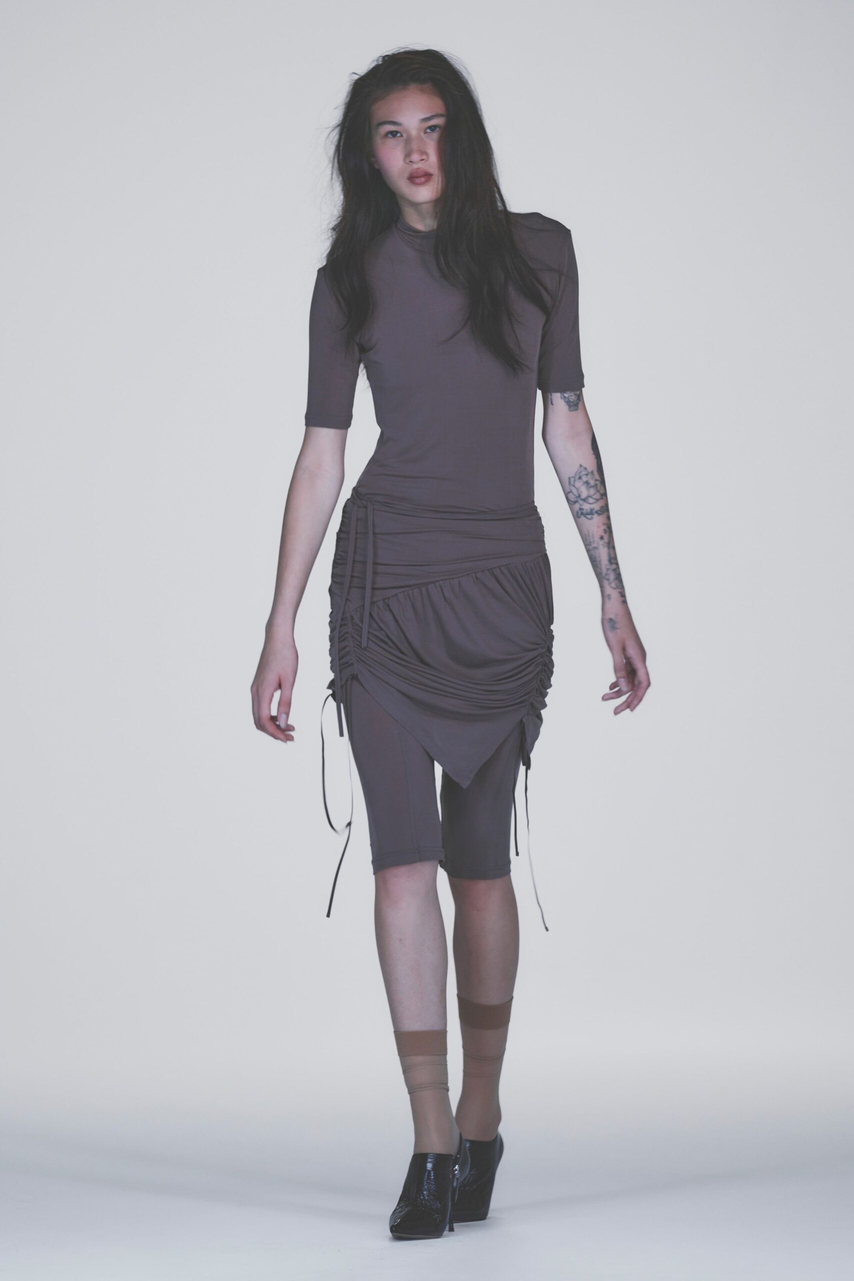 Smoke-brown stretch jersey top with a snug mock neck. Ruched asymmetrical skirt, drawcord ends hanging past the knee. Matching jersey cycling shorts visible beneath the pointed hem. Sheer beige ankle socks. Black patent booties with a side zip and knife heel. Like leaving a warehouse casting call into cold early-evening drizzle.