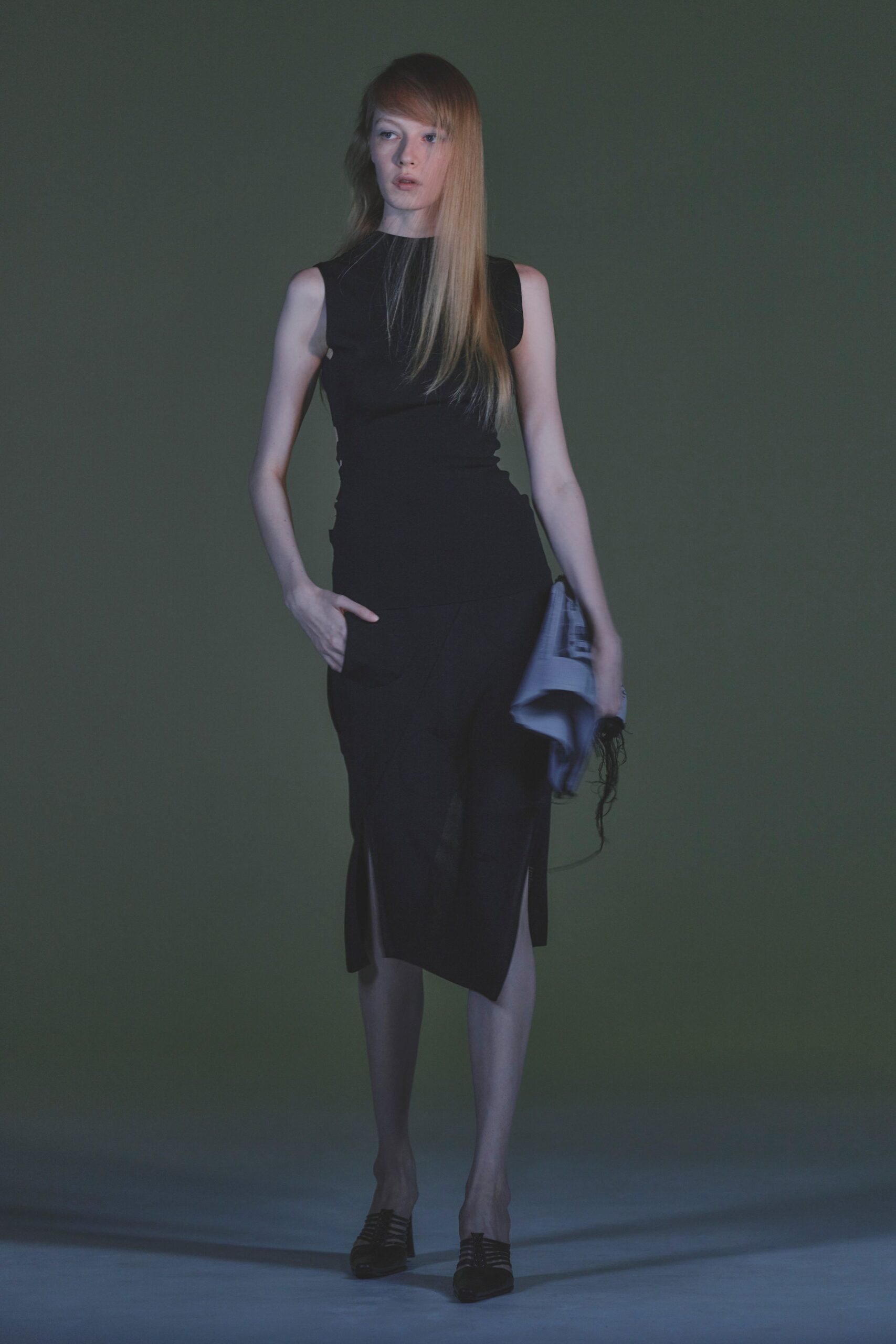 Black sleeveless jersey top, ruched tight through the waist. Black midi skirt with an off-center slit and scooped hip pocket. Pale printed scarf clutched in one hand, black fringe trailing. Black cage-front mules. Walking home from the office with the folded paper still in hand.
