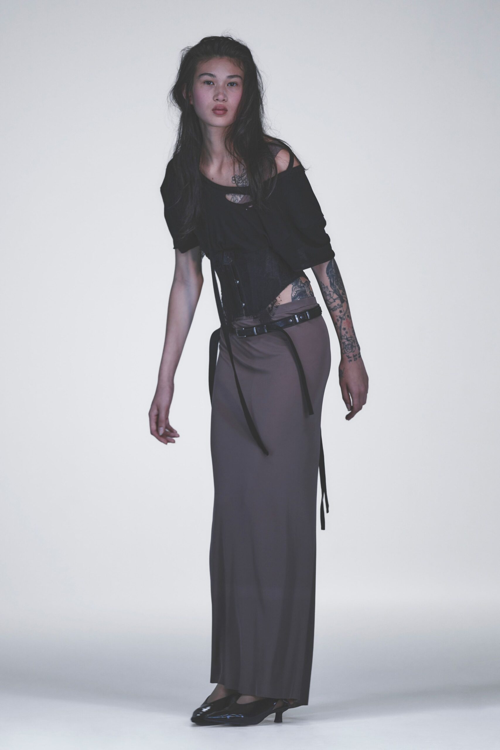 Black jersey top slipping off one shoulder, cut with a chest keyhole. Black corset panel with hook closures and dangling ribbon ends. Taupe column skirt in fluid knit, split at the side seam. Low black double belt with silver grommets. Patent kitten heels. Like leaving a tattoo appointment just before the rain starts.