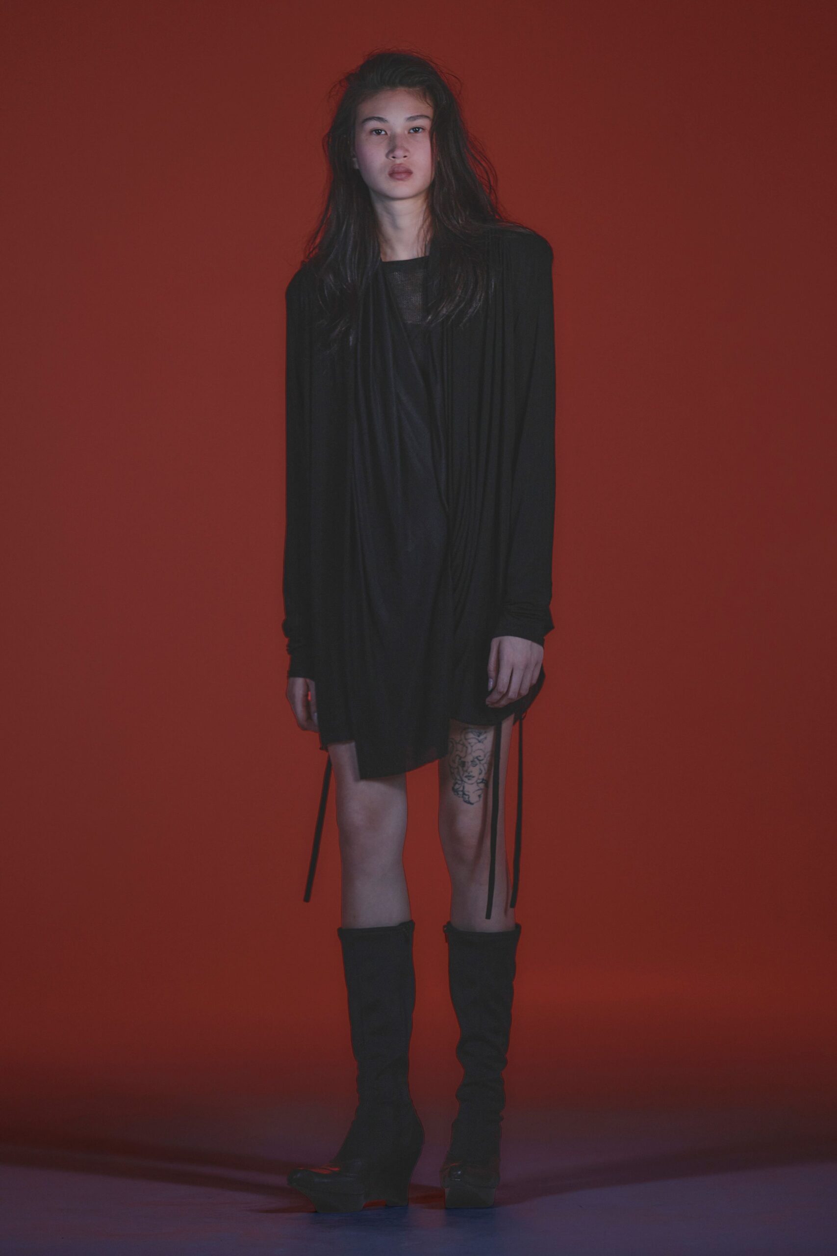 Black draped jersey mini dress over a sheer mesh crewneck, the hem falling longer on one side. Long black cardigan with ribbon ties hanging past the knees. Slouchy suede boots. In a rehearsal room after midnight, radiator hissing.