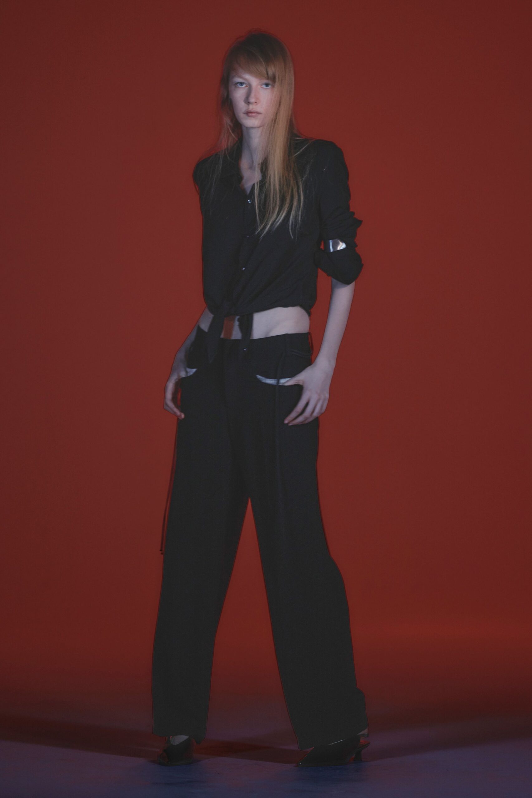 Black shirt knotted above the navel, sleeves pushed to the elbow. Mirror-bright metal cuff strapped over one sleeve. Low-slung black wide-legs with curved cutouts flashing white pocket linings. Pointed black mules with a small flare heel. Waiting under the red exit sign after the club lights came up.