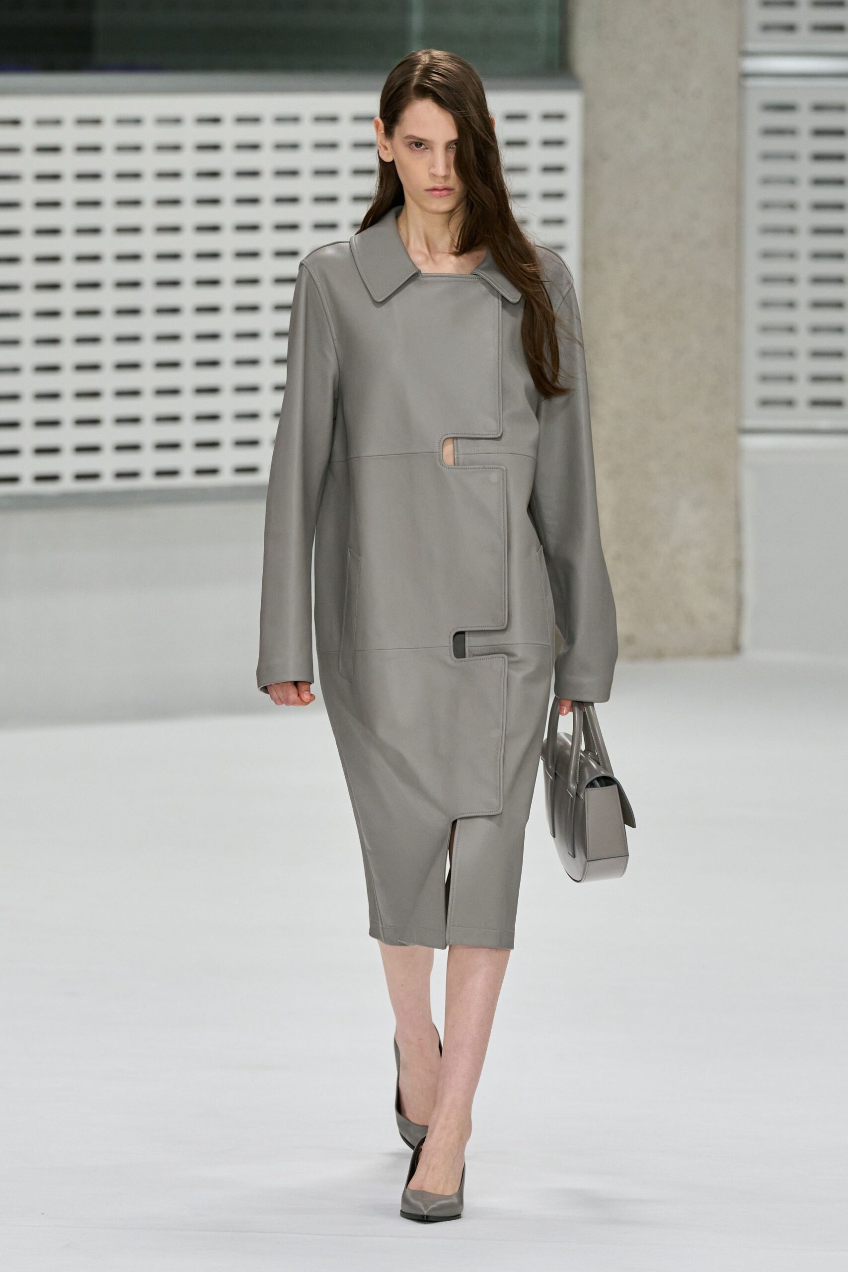 Dove-grey leather sheath with puzzle-piece panels and two narrow cutouts. Matching structured tote, flap standing off the side. Grey leather pumps with a gently squared toe. Early train to the finance district, carrying papers nobody else should read.