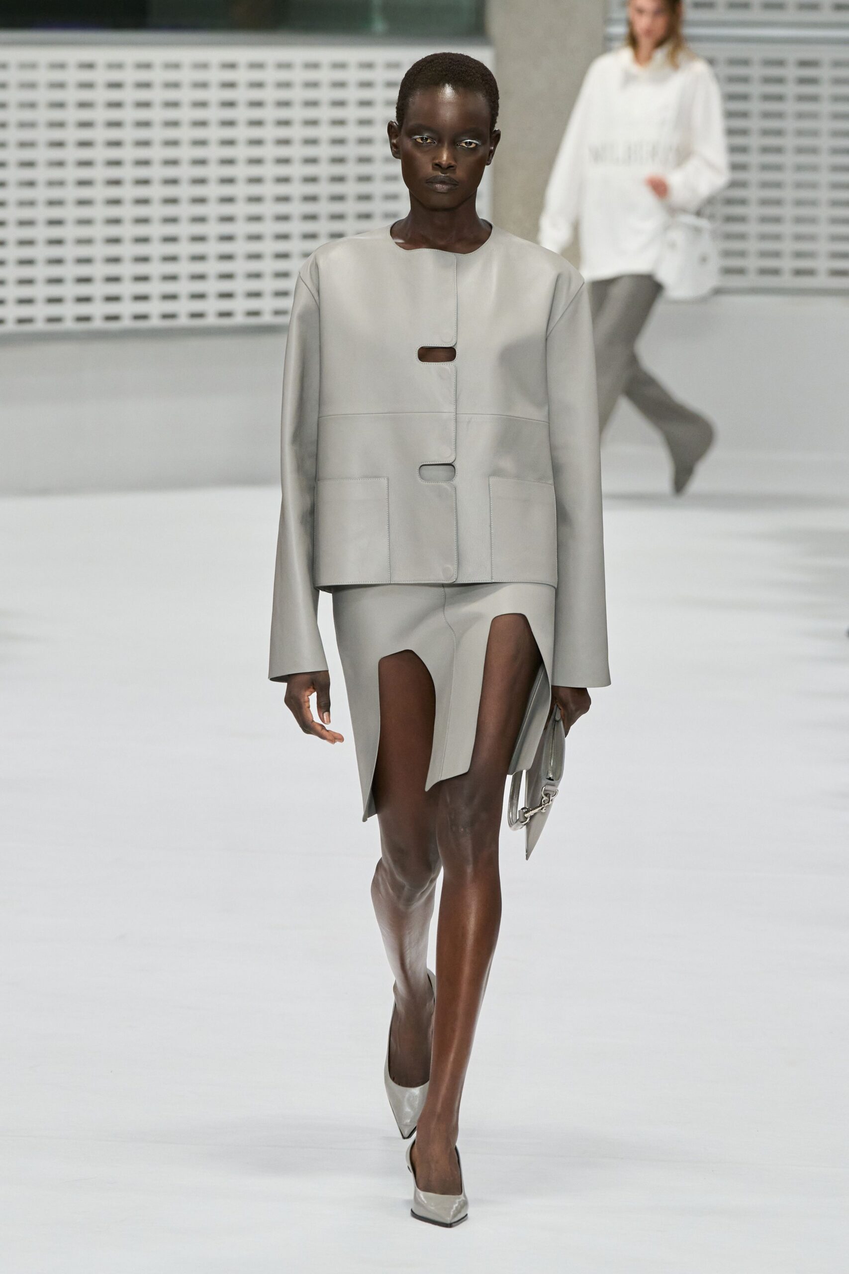 Pale dove leather jacket with pill-shaped cutout closures and square patch pockets. Matching leather mini with two deep front slits curving around the thighs. Slim leather clutch with a short silver curb-chain strap. Pointed dove leather pumps with a sharply squared vamp. Crossing a hushed museum atrium before the doors unlock.