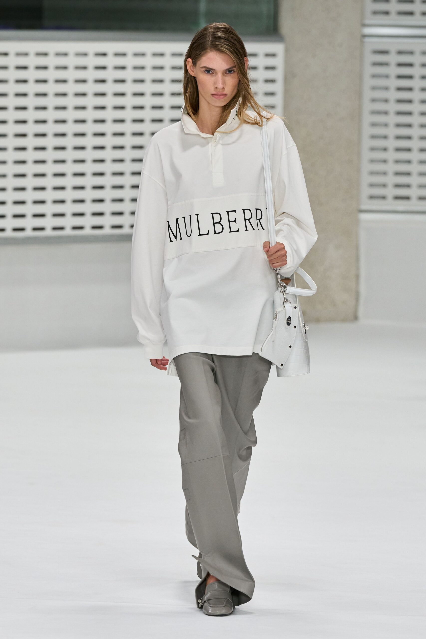 Oversized white polo in smooth jersey, tall collar tipped open. Pearl-grey trousers with pressed seams and a front hem slit. White croc-embossed shoulder bag with silver postman-lock hardware. Grey leather loafers, squared off at the toe. Walking through a quiet office atrium before anyone else arrives.