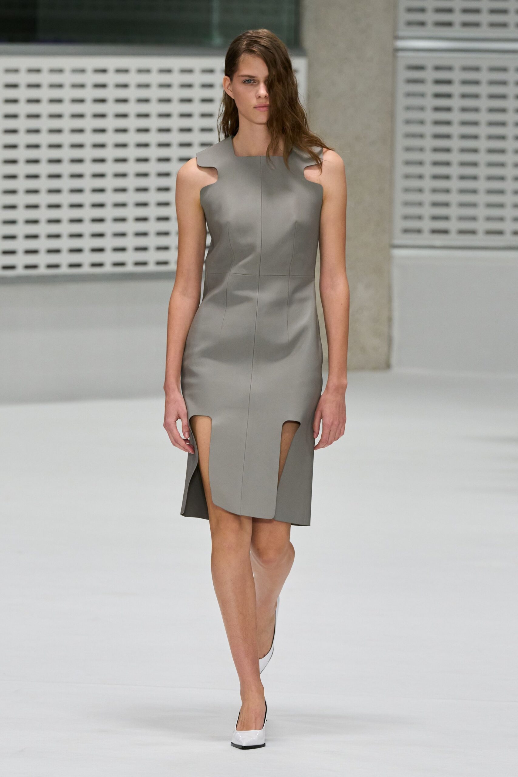 Matte dove-grey leather sheath, scalloped arm openings and two thigh cutouts. White square-toe pumps with black sole edging. Crossing a museum atrium before the building opens to the public.