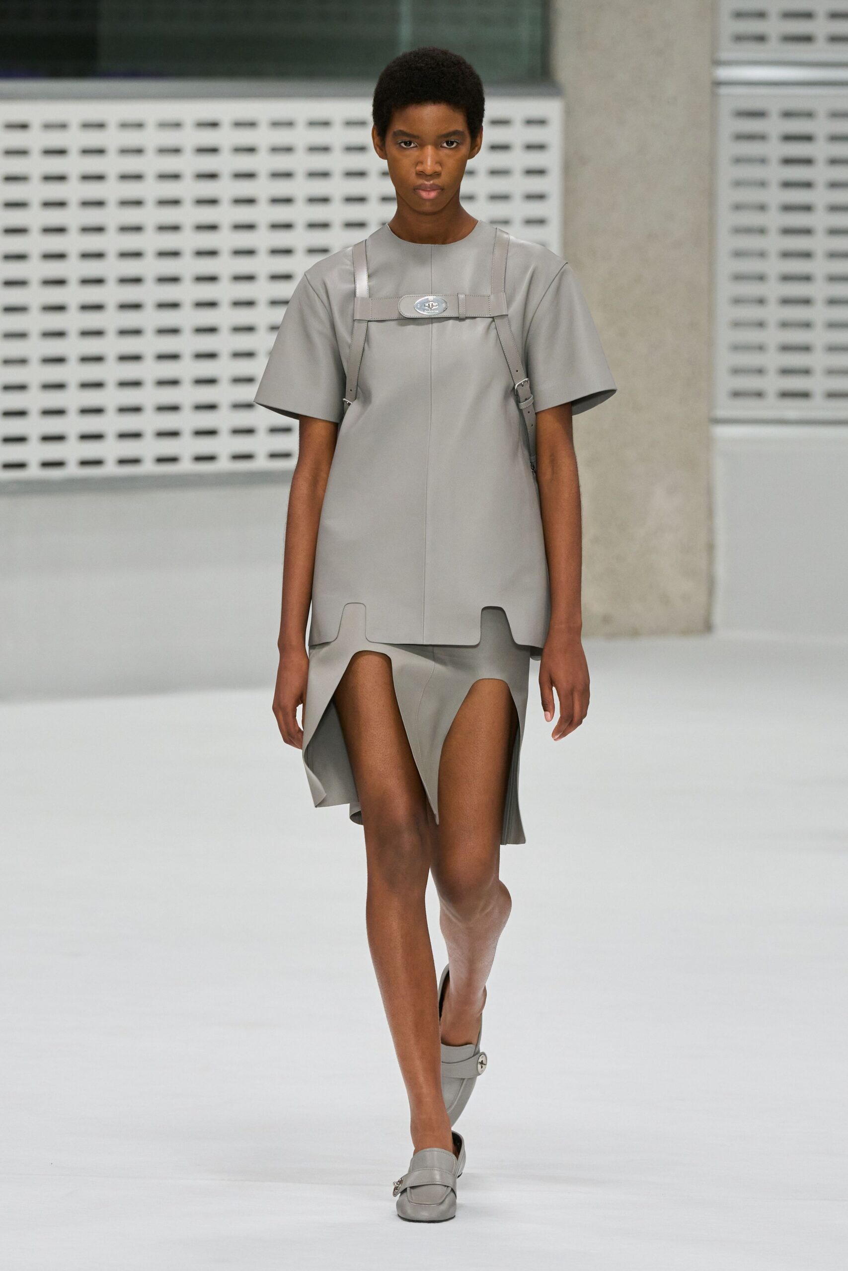 Pale grey bonded tunic with a central seam and squared notched hem. Matching leather chest harness, oval plaque buckled across the sternum. Asymmetric skirt cut into deep inverted arches that bare the thighs. Soft grey loafers with a strap and small silver horsebit. Museum atrium hush, footsteps carrying across the stone floor.