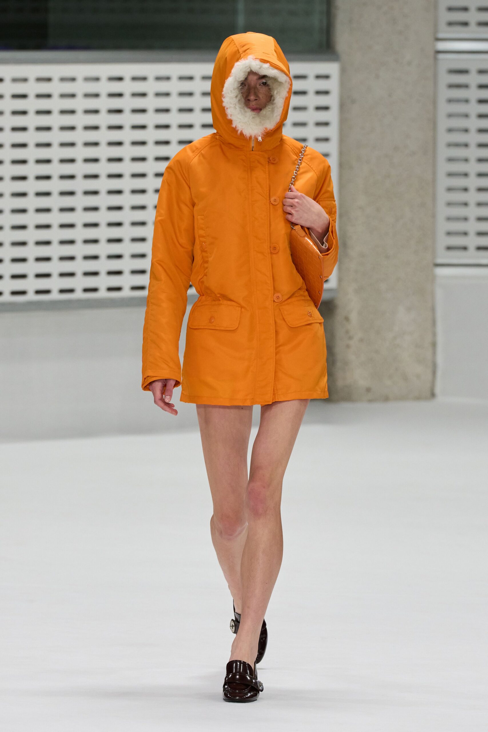 Tangerine shell parka, hood ringed in dense cream shearling. Small tangerine crocodile-embossed shoulder bag on a silver chain. Dark patent loafers with a bright metal horsebit. Crossing a ferry deck in March, wind needling through the railings.