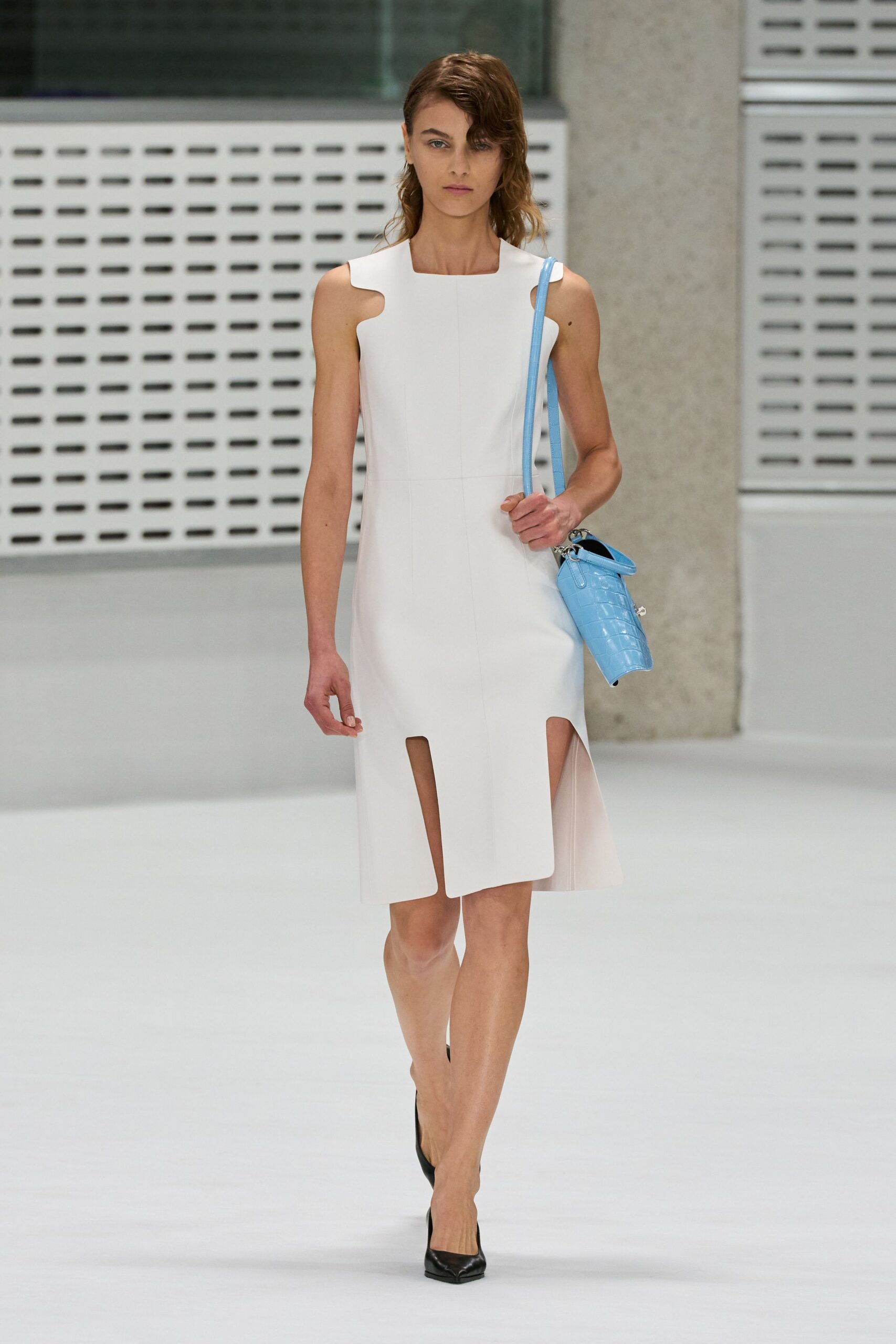 White sleeveless sheath dress with cutout shoulders and two long front hem slits. Powder-blue crocodile-embossed bucket bag with a silver ring at the handle. Black pointed pumps. Crossing a museum atrium before the doors open to the public.