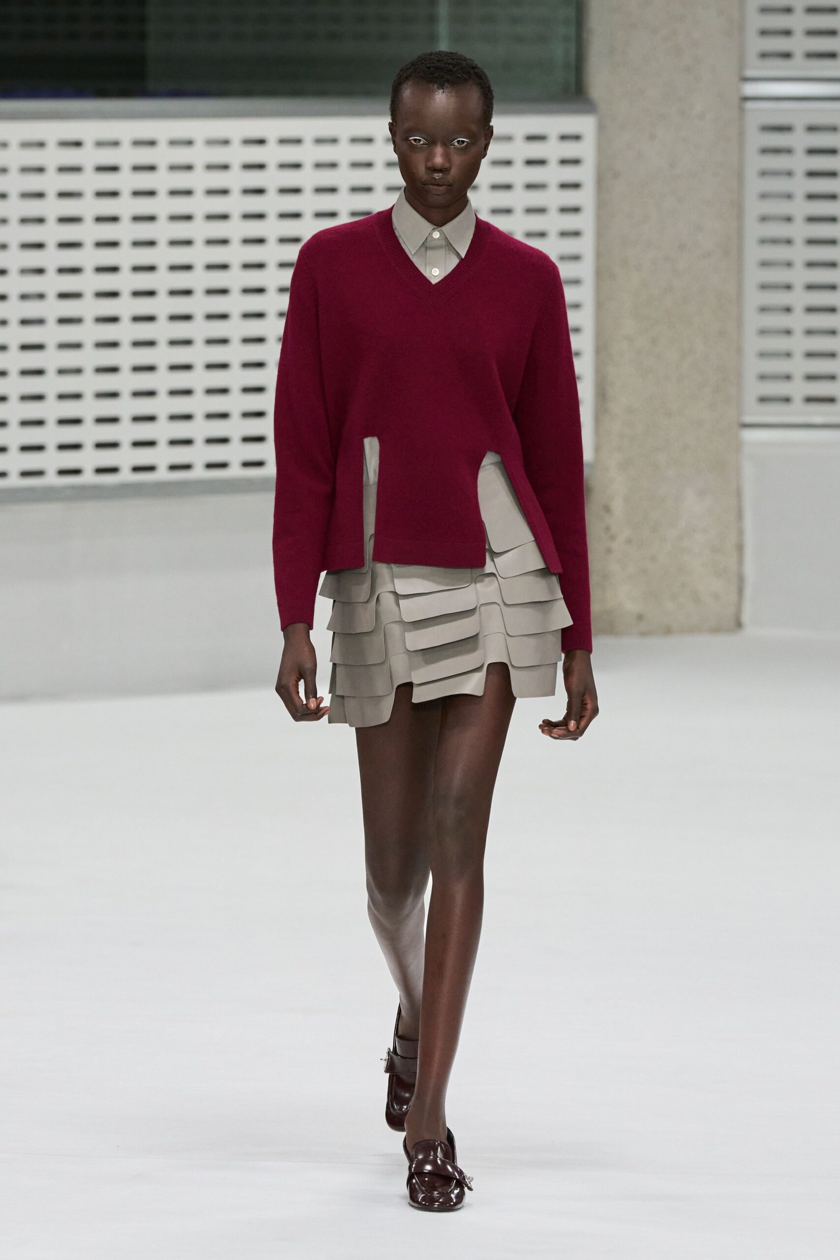 Burgundy V-neck knit with two deep front hem slits. Pale grey poplin collar peeking cleanly above the neckline. Stone mini built from stacked rounded flaps, one lifting at center. Oxblood patent loafers with a small silver bit. Crossing a quiet campus courtyard before the library opens.