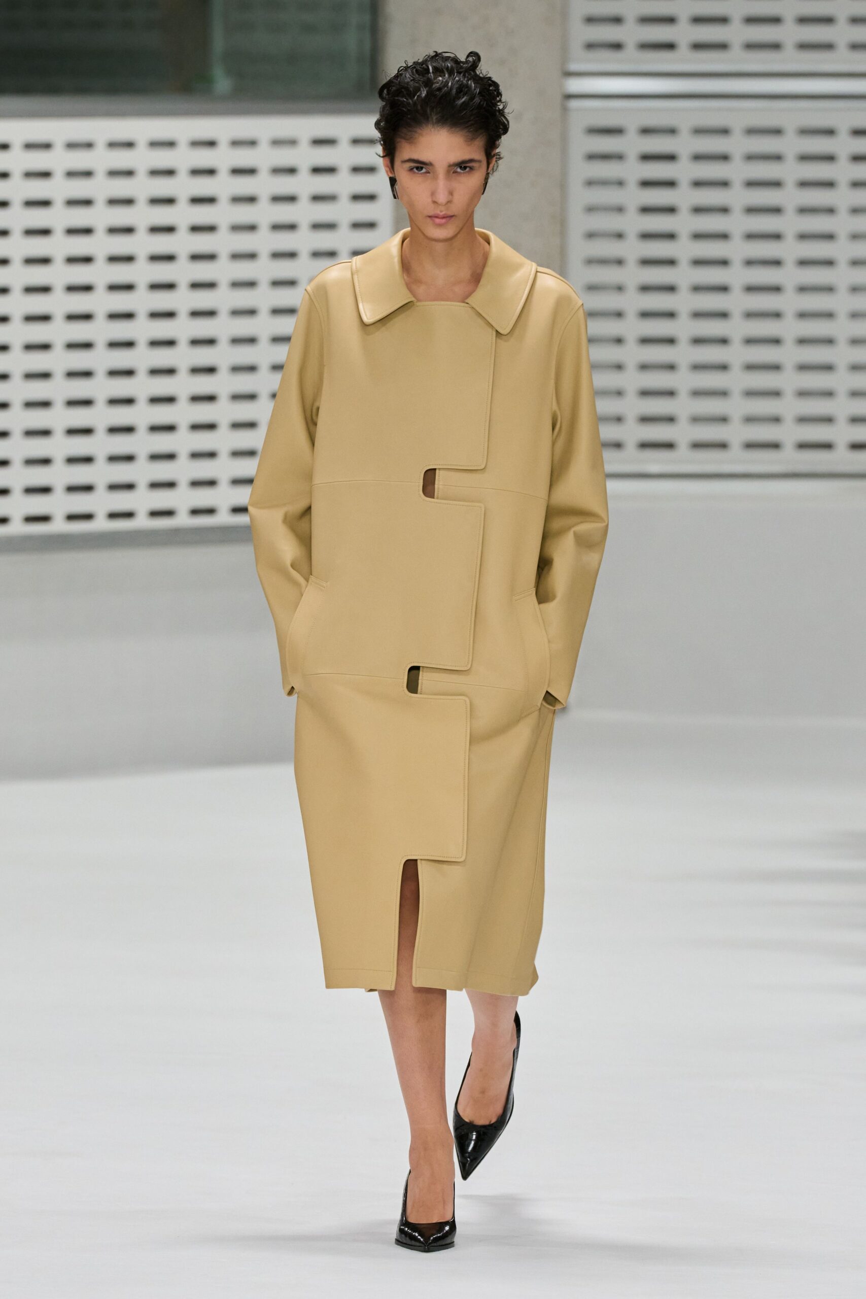 Buttercream leather coat with a stepped front panel cut into keyhole notches. Matching knee-length skirt split by the same puzzle-edged overlap. Black patent pumps, knife-point toes. Municipal atrium at noon, carrying plans nobody else has seen yet.