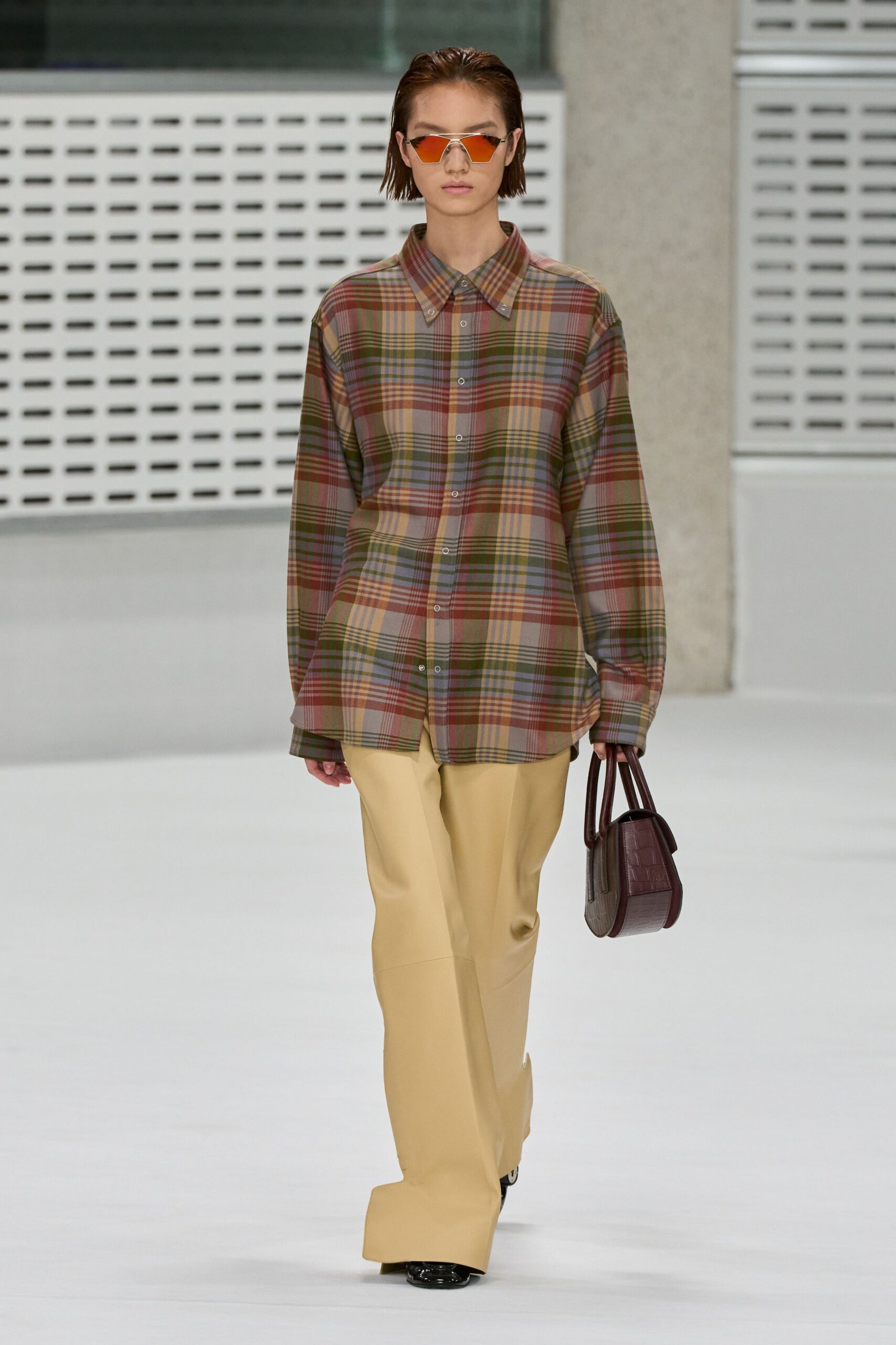 Angular silver frames with orange mirrored lenses. Oversized madras plaid shirt, button-down collar sitting flat and wide. Buttercream trousers with a long break pooling over the shoe. Burgundy crocodile-embossed top-handle bag, stiff and boxy. Walking into the office park after lunch with sun still glaring off the glass.