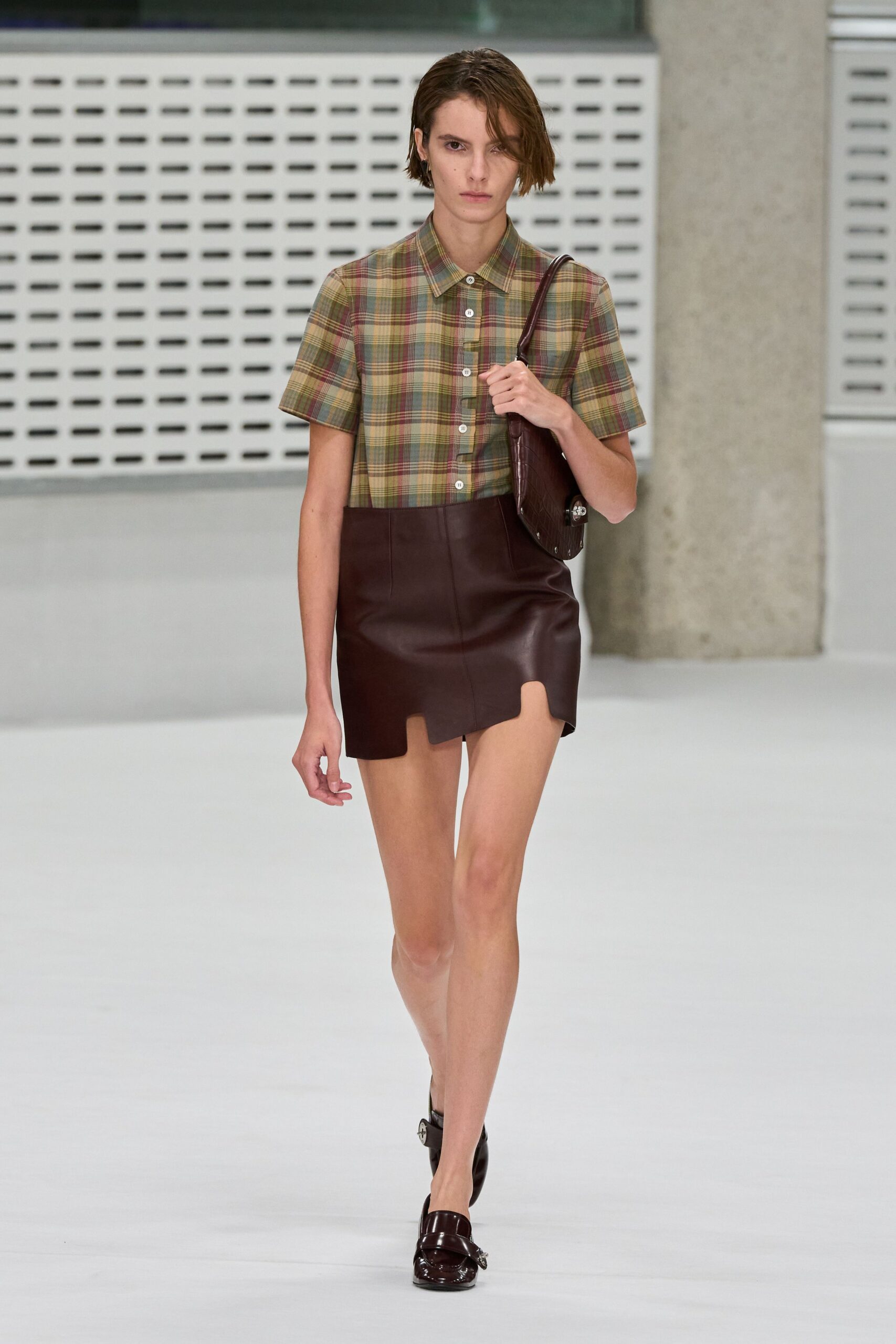 Muted tartan short-sleeve shirt, white buttons marching down the center. Oxblood leather mini with a scalloped cutout hem at one thigh. Embossed oxblood flap bag tucked tight under the arm. Glossy oxblood loafers with silver horsebit hardware. Walking across campus after rain, library books knocking in the bag.