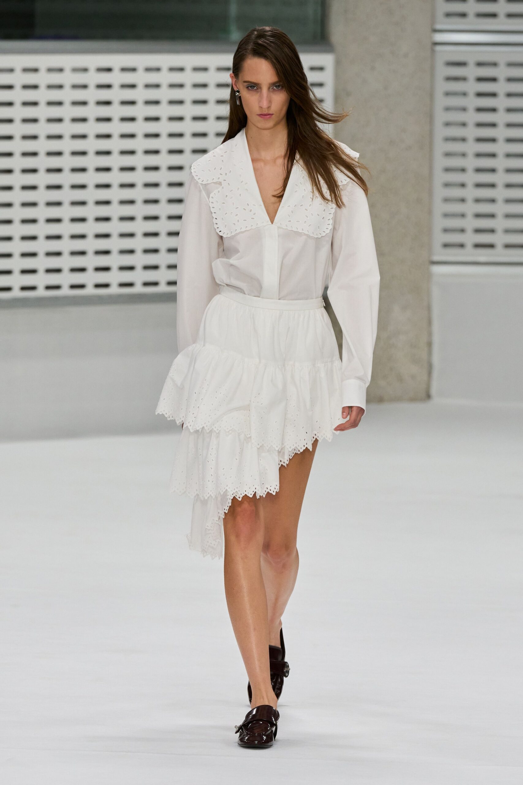 White cotton blouse with a broad broderie anglaise sailor collar. Tiered white skirt, eyelet ruffles dropping into an uneven handkerchief hem. Dark oxblood loafers with a strap across the square toe. Walking from a registry office into wind off the river.