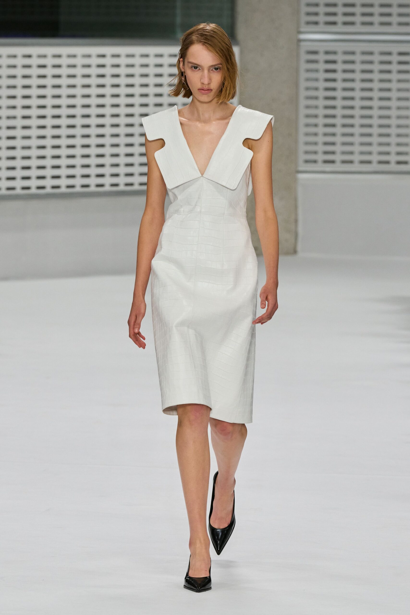 Ivory croc-embossed sheath dress with a deep V and squared wing collar. Black patent pumps with sharply pinched toes. Crossing a near-empty civic atrium before the offices unlock.