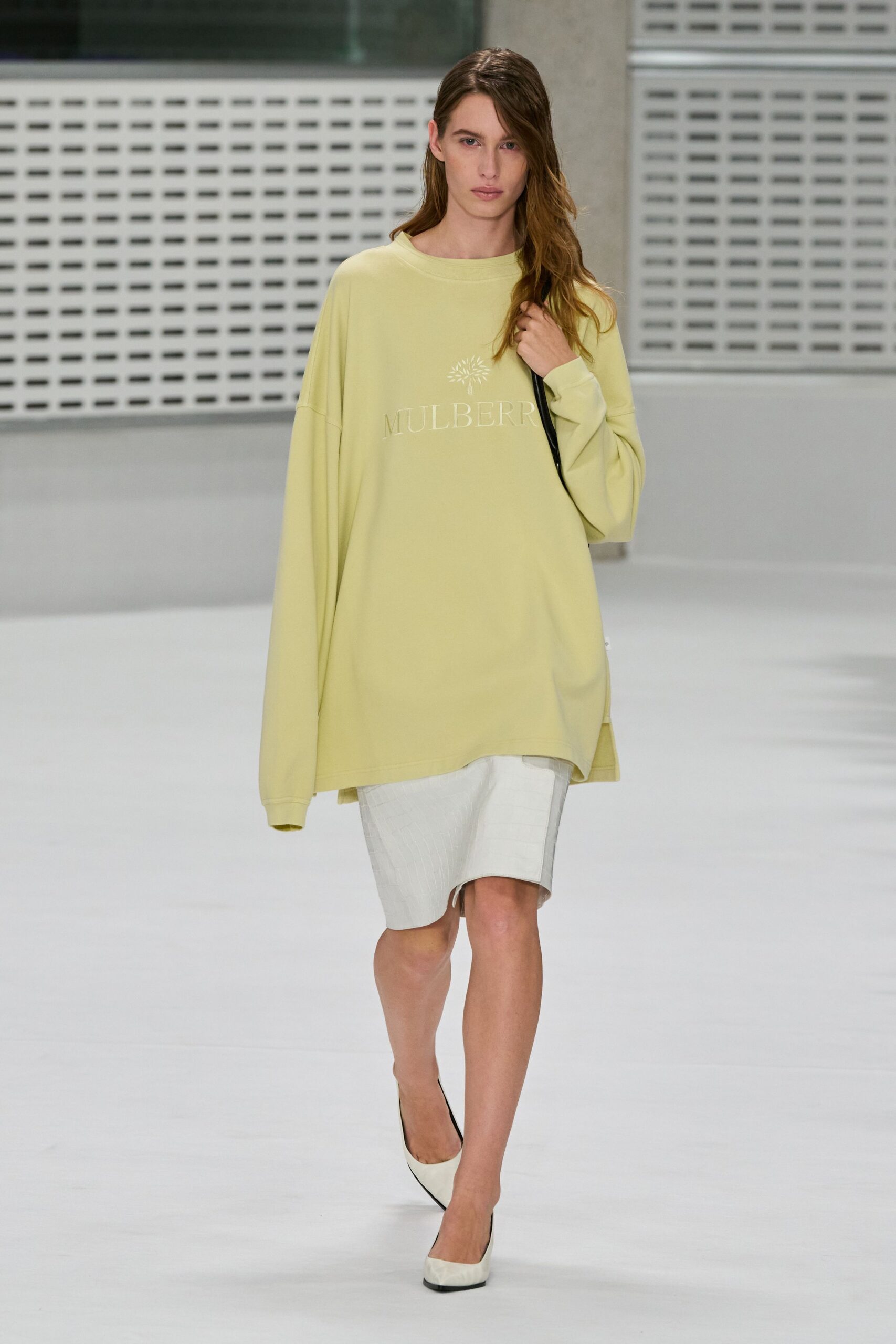 Oversized butter-yellow sweatshirt, flocked chest logo and sleeves swallowing the hands. Pale knee-length shorts with faint crocodile embossing and a dangling drawcord tip. Cream pumps with sharp black-edged soles. Sunday airport lounge, coffee gone cold before boarding starts.