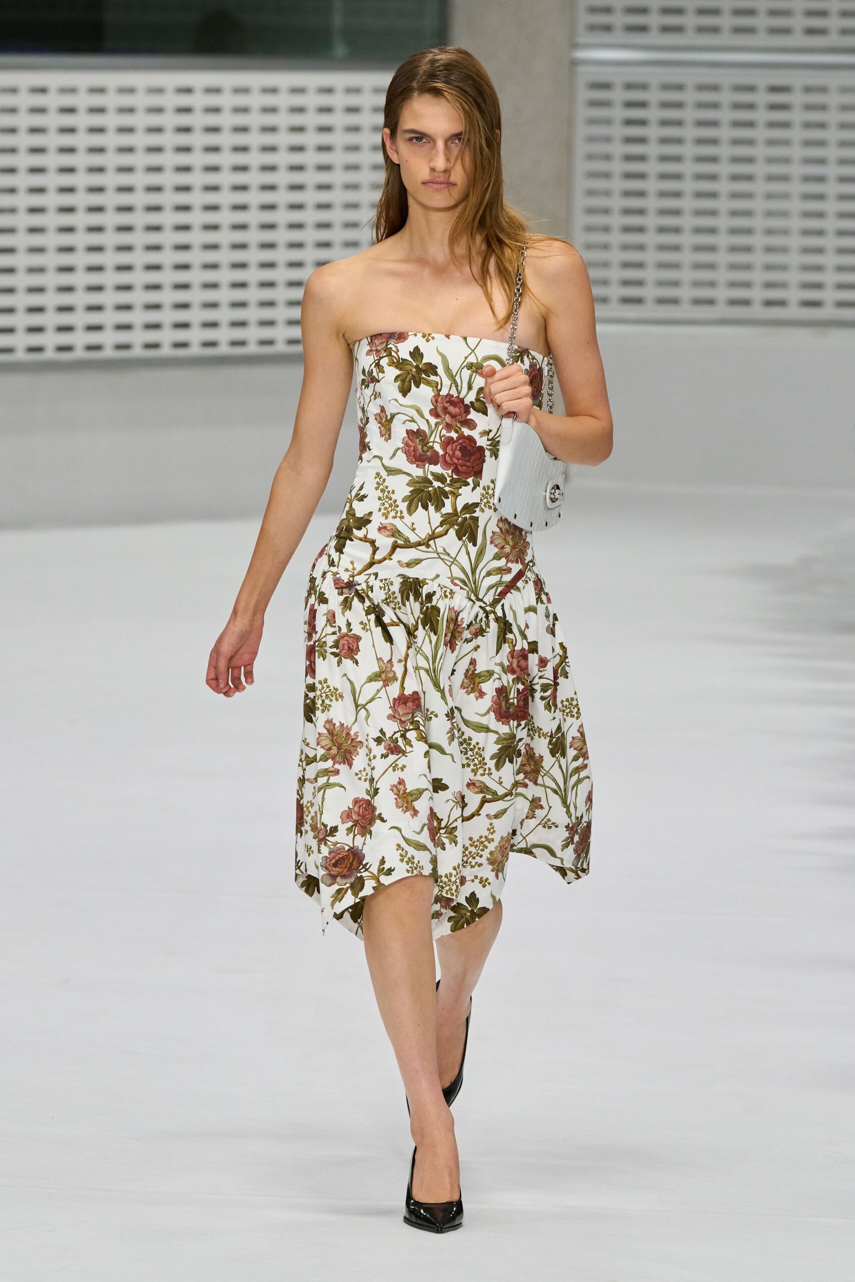 Strapless white dress printed with dusty roses and olive trailing vines, skirt dropped into soft gathers. White crocodile-embossed shoulder bag on a silver chain strap. Black patent pumps with a narrow pointed toe. Hotel breakfast after the wedding, still carrying last night's flowers.
