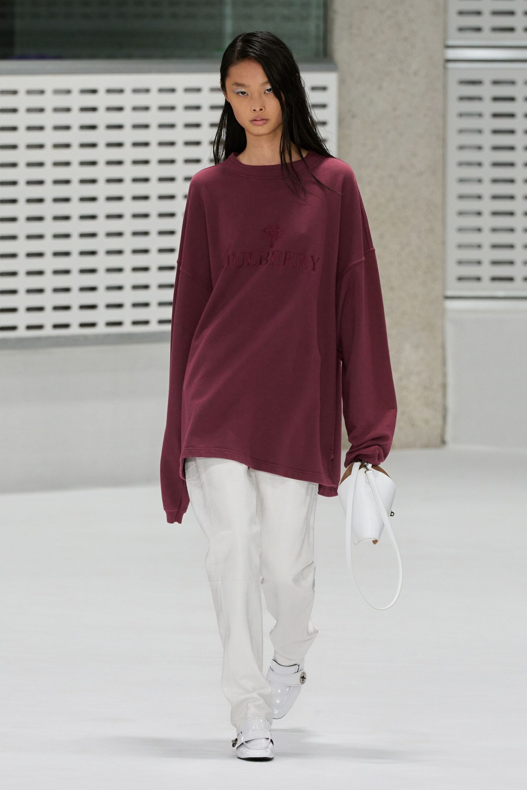 Oversized mulberry crewneck sweatshirt with tonal embossed lettering across the chest. White straight-leg trousers in crisp twill, creased through the front. White leather pouch bag on a stiff loop handle. White patent flats with buckled straps at the vamp. Early train to the airport, coffee cooling in a paper cup.