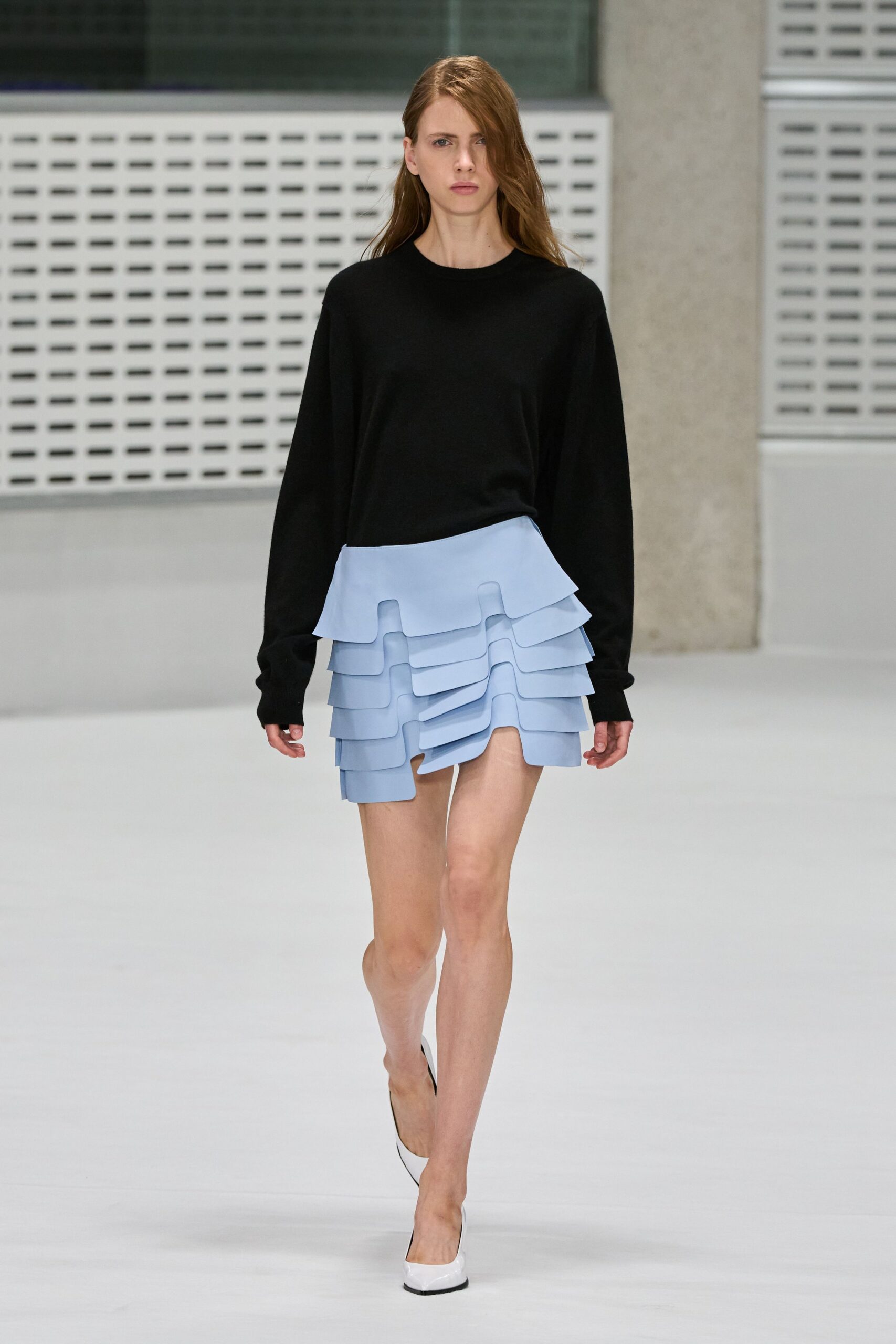 Black fine-gauge sweater hanging long through the sleeves. Powder-blue mini built from stacked rounded flanges, one panel skewed at center. White pumps with black-edged soles. Crossing an office atrium before the building fully wakes up.