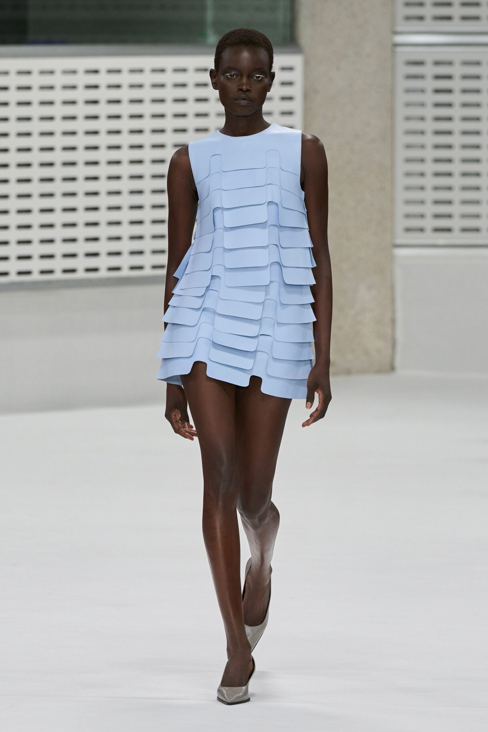 Powder-blue sleeveless mini built from stacked rounded flaps, hem dipping unevenly. Silver leather pumps with a sharply squared toe. Museum atrium at noon, light bouncing off every pale surface.