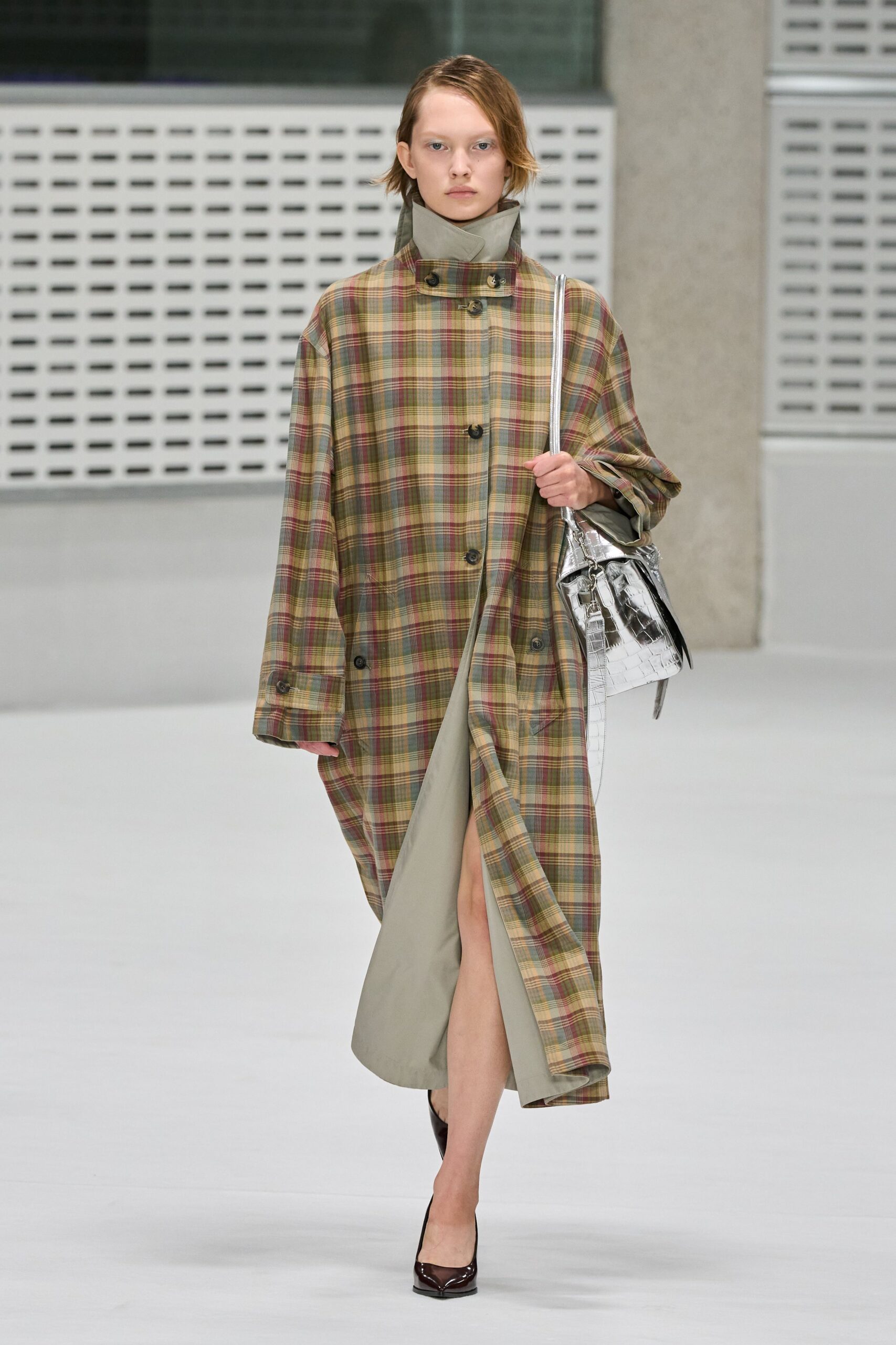 Oversized muted plaid coat, storm collar buckled high over a sage stand neck. Pale sage column dress opening to a sharp front slit. Mirror-silver crocodile-embossed shoulder bag with a long rigid strap. Oxblood patent pumps with squared toes. Walking into a civic building with rain still drying on the steps.
