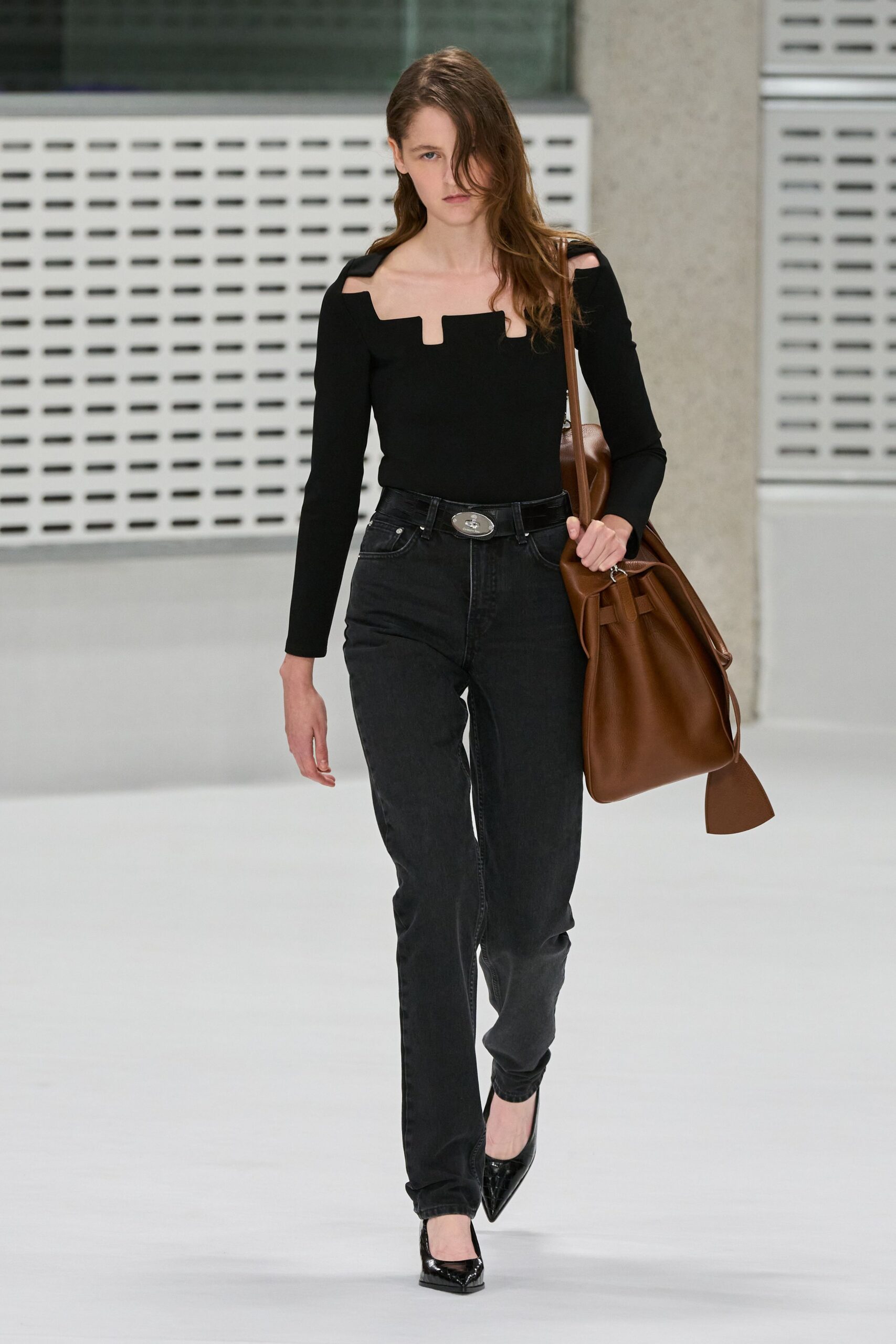 Black knit top with a squared notch neckline and cutout shoulders. Washed black jeans, high-waisted with a narrow straight leg. Oval silver buckle at the waistband. Cognac leather bucket bag with a long tapered tie end. Black patent pumps with sharp pointed toes. For crossing a corporate plaza after missing the car service.