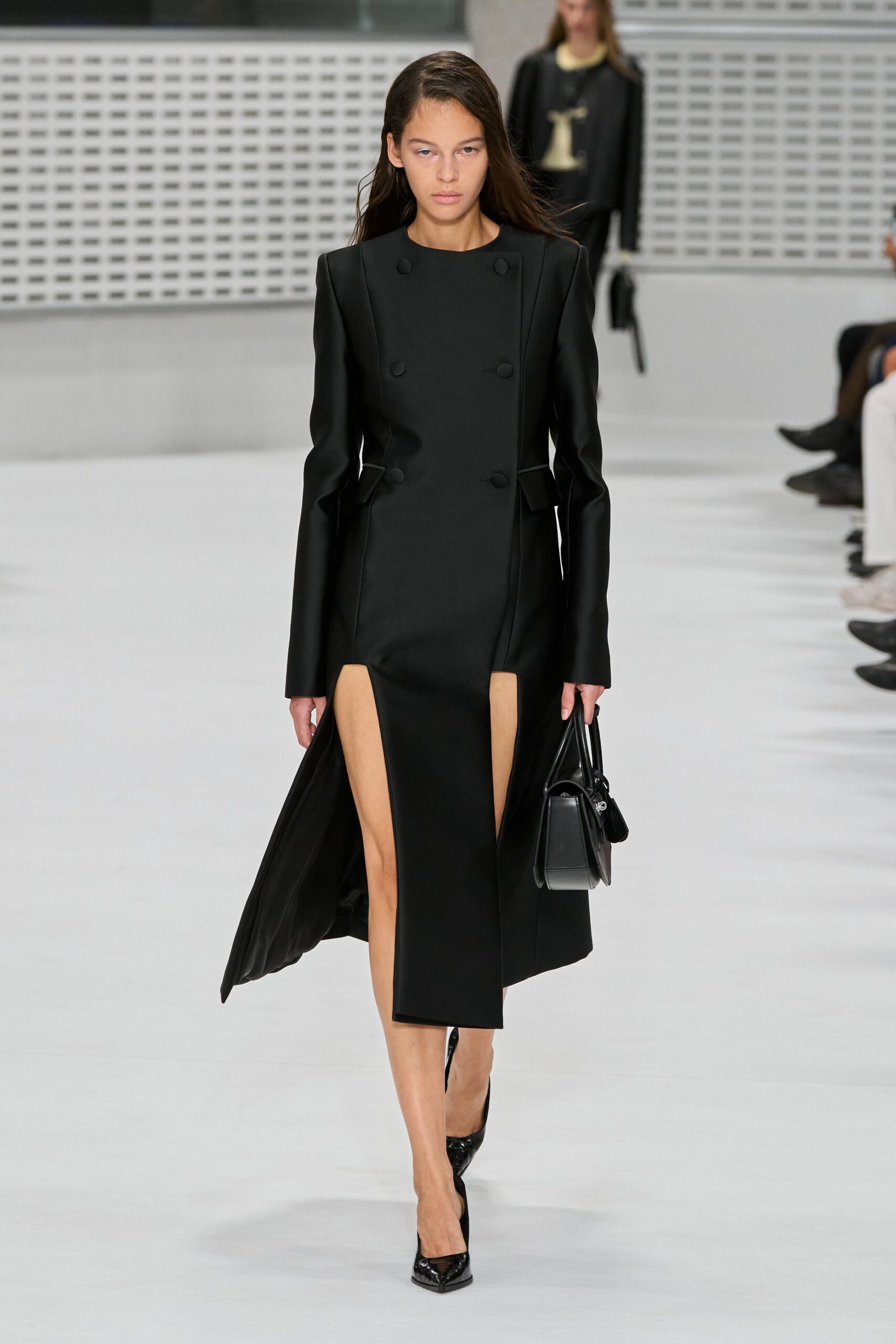 Black tailored dress coat with padded shoulders and six covered buttons. Boxy black top-handle bag with a tiny zip case clipped on. Black patent pumps, sharply pointed at the toe. For the courthouse steps after rain, file tucked under one arm.