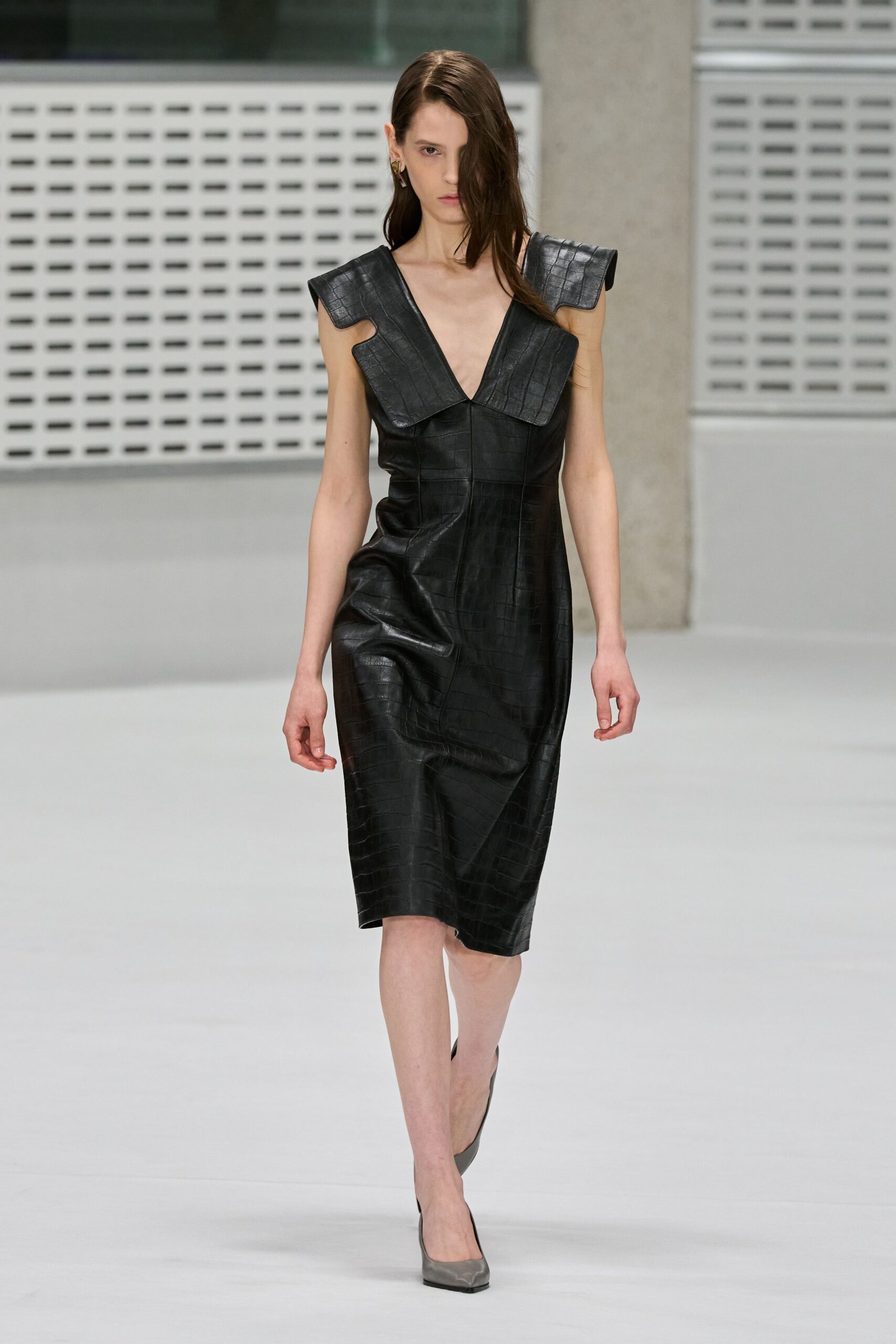 Small gold hoop tucked under wet hair. Black crocodile-embossed leather dress with huge folded shoulder flanges and a plunging V. Grey pointed pumps. Walking into a concrete office atrium after the rain stopped.