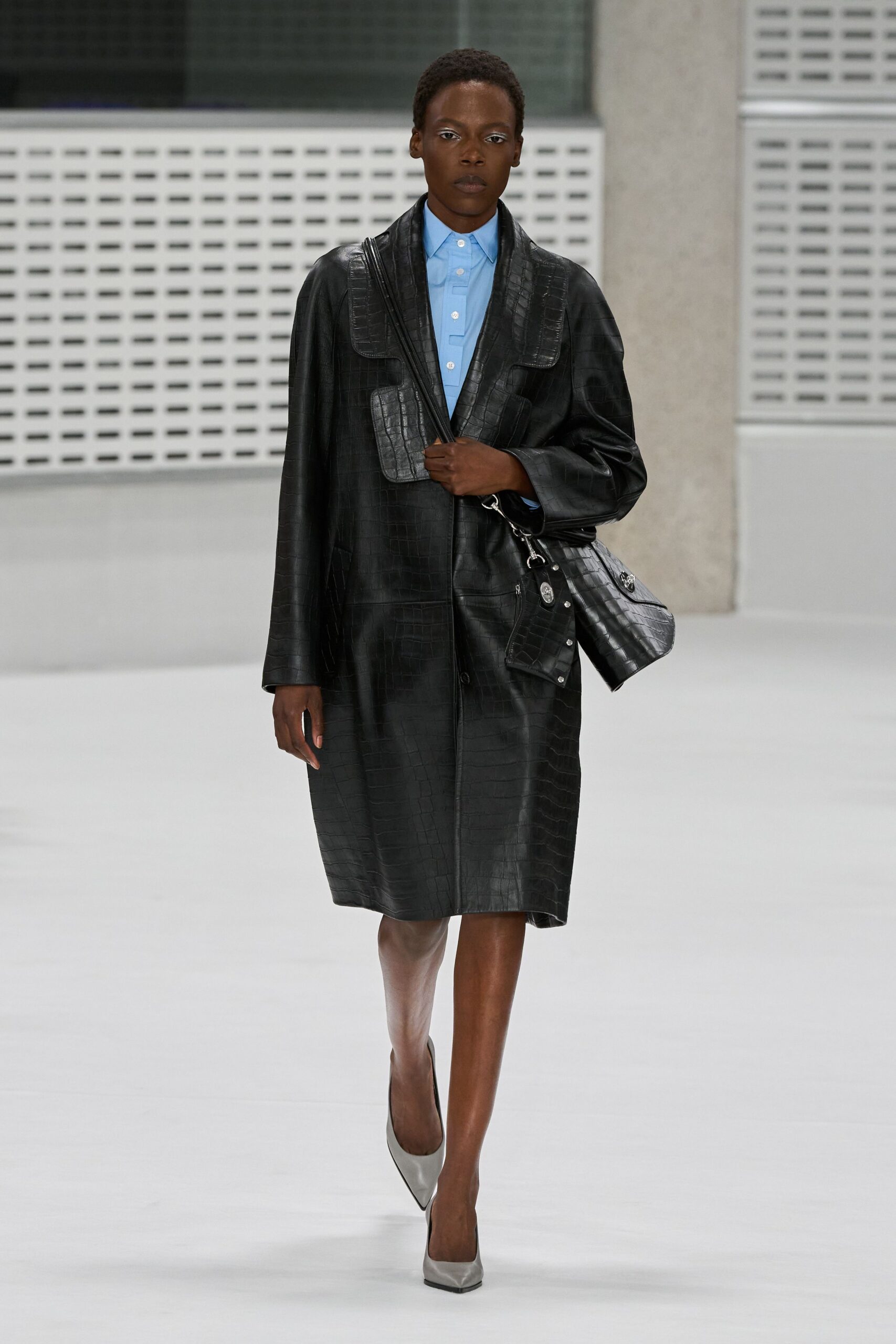 Black crocodile-embossed leather coat, cut long with oversized flap pockets. Powder-blue cotton polo buttoned tight to the throat. Matching crocodile-embossed bag with a clipped silver chain strap. Pewter pointed pumps, dark sole edging visible at the tip. Walking into a municipal building with rain still drying on the steps.