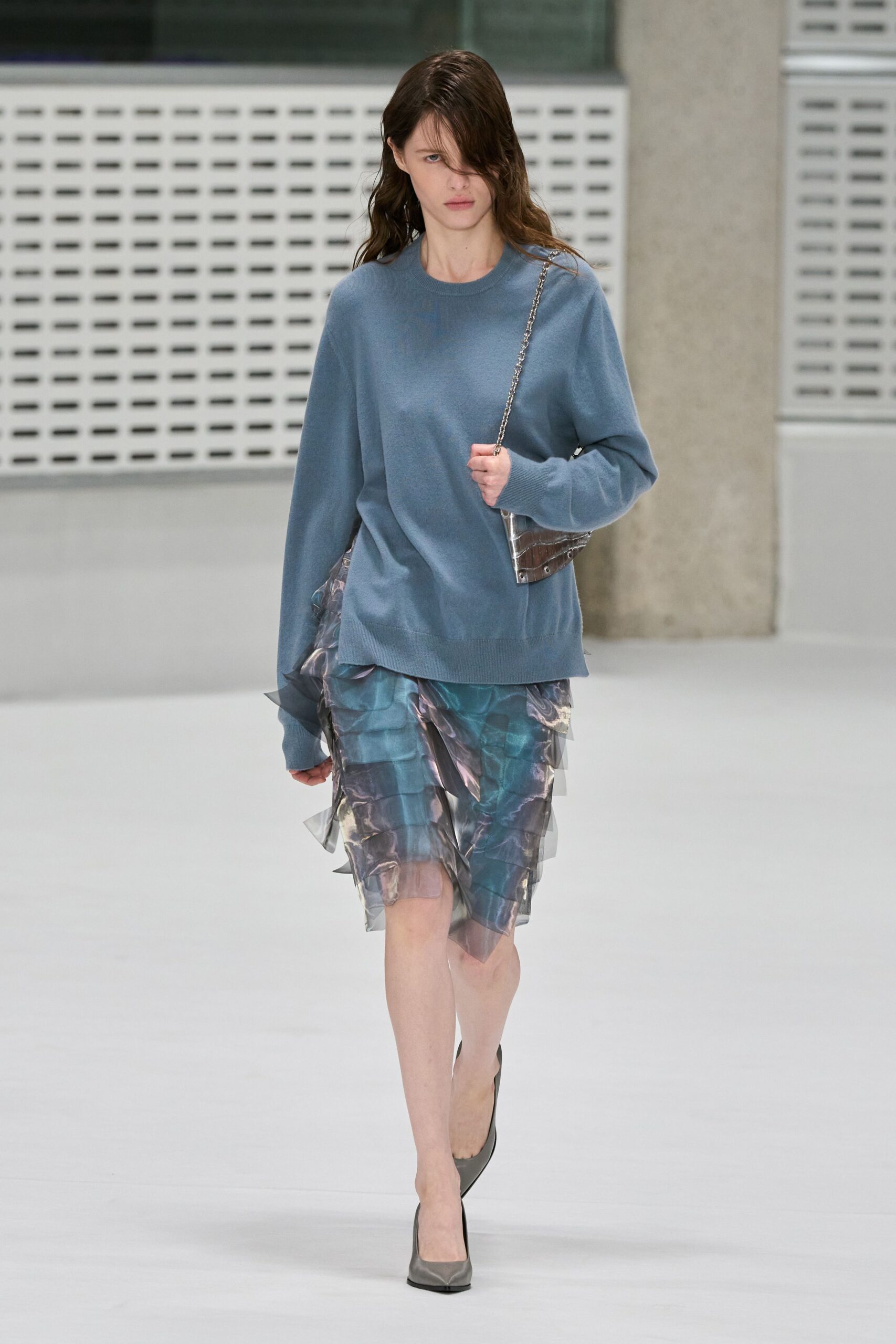 Dusty blue fine-gauge sweater, cuffs long enough to swallow the knuckles. Silver chain strap on a crinkled metallic pouch. Blue-grey printed skirt under sheer organza ruffles, hem tipping into points. Steel grey leather pumps. Office atrium on a wet Monday, carrying yesterday into the meeting.