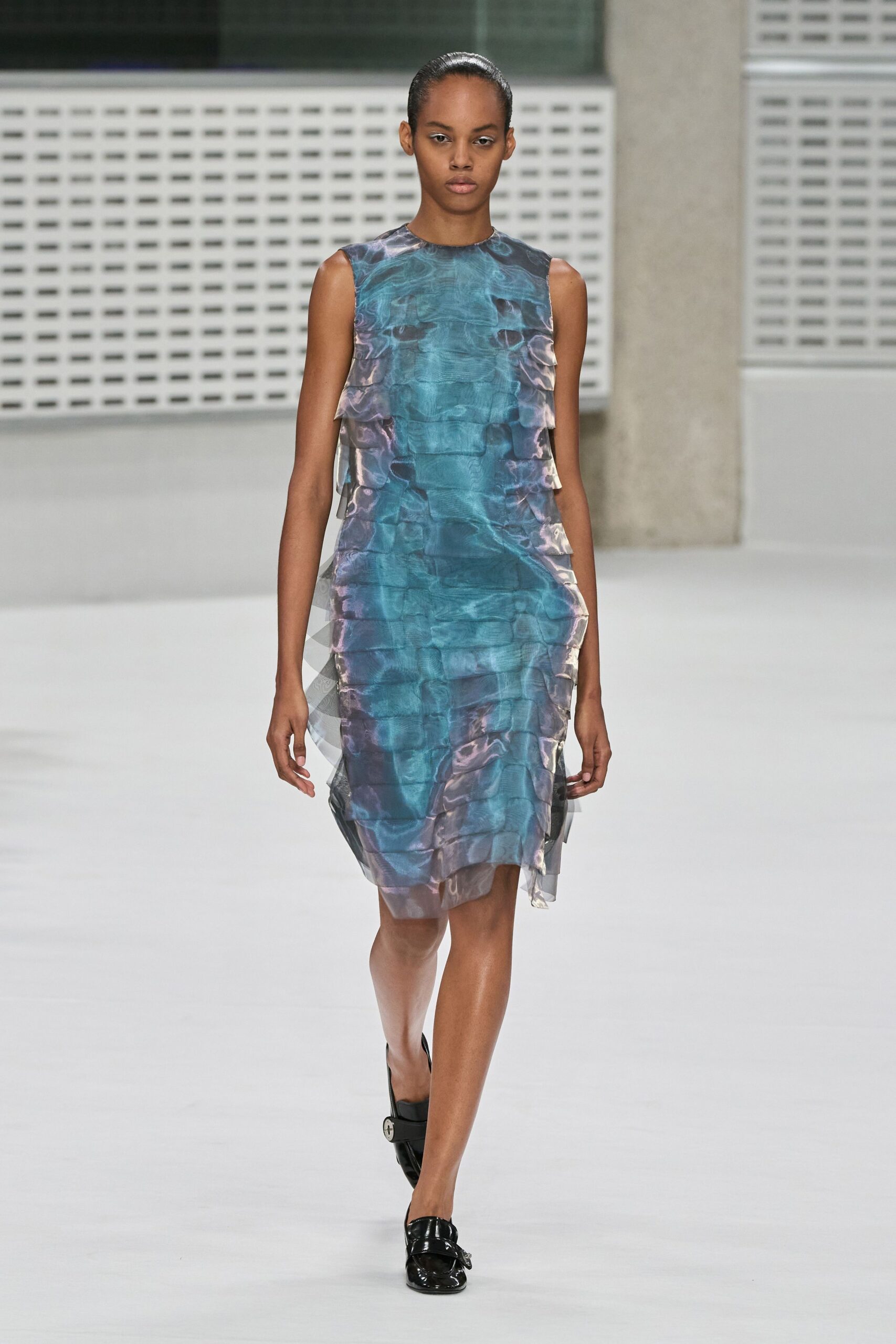 Sleeveless iridescent organza dress in watery teal and lilac, paneled into translucent side flounces. Black patent loafers with a silver strap buckle at the vamp. Museum atrium at noon, rain still drying on the steps outside.
